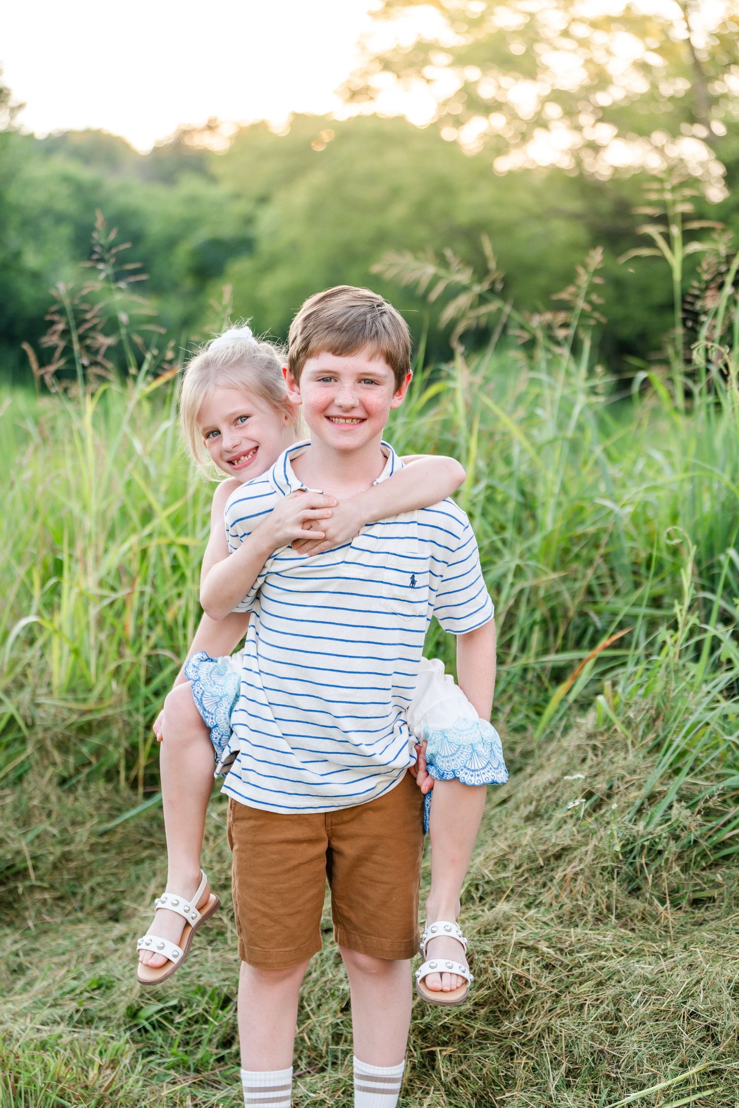 Boy giving younger girl a piggyback ride in a sunny green field at golden hour.
