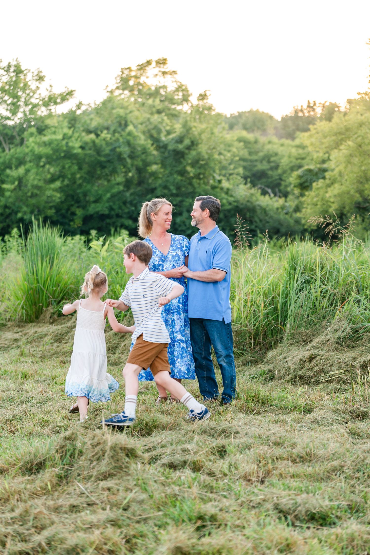 A happy family of four enjoys a sunny outdoor stroll through a lush green meadow, with parents and children dressed in casual summer attire.