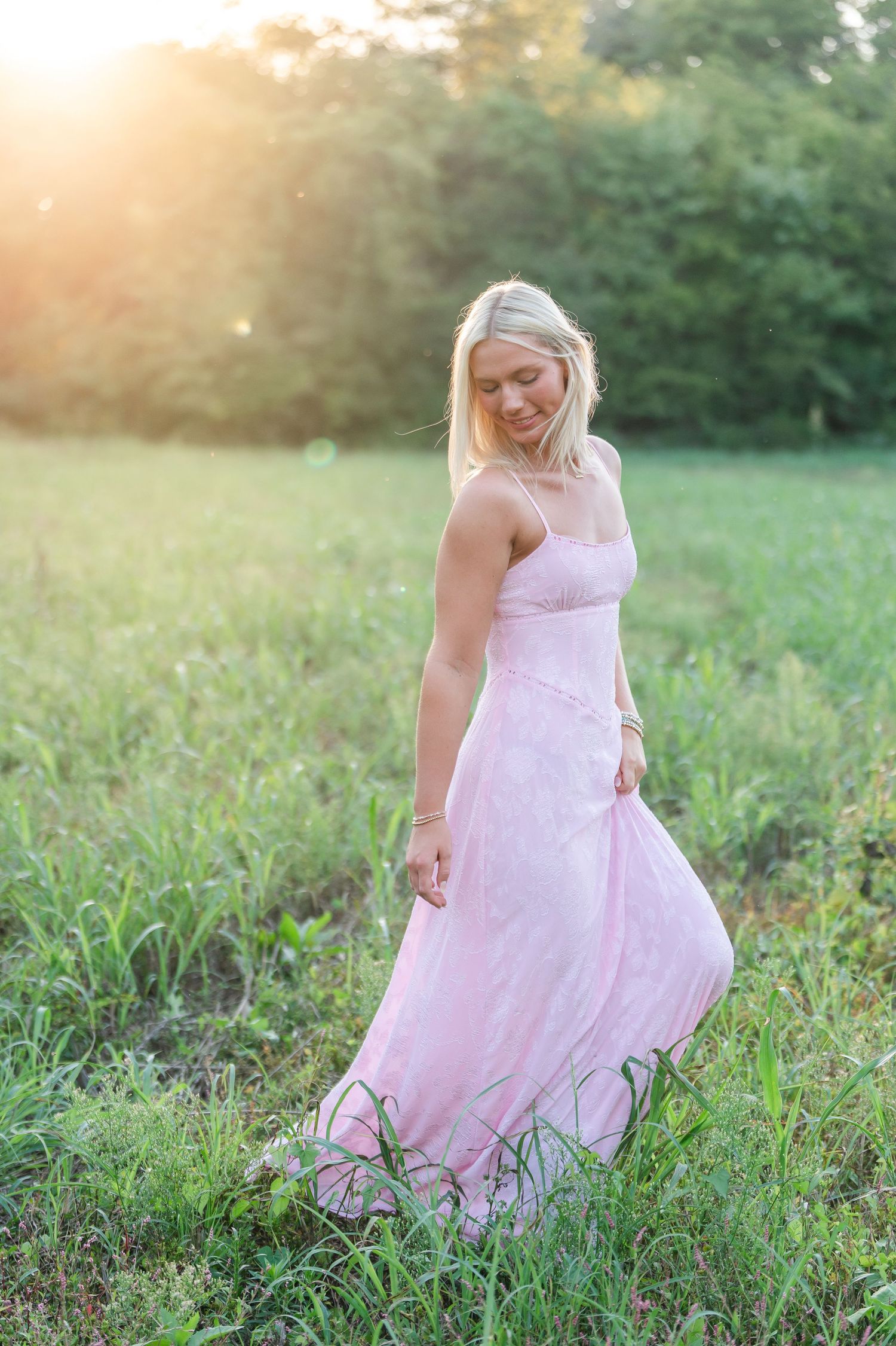 Blonde woman in pink maxi dress walking through green field bathed in golden sunset light.