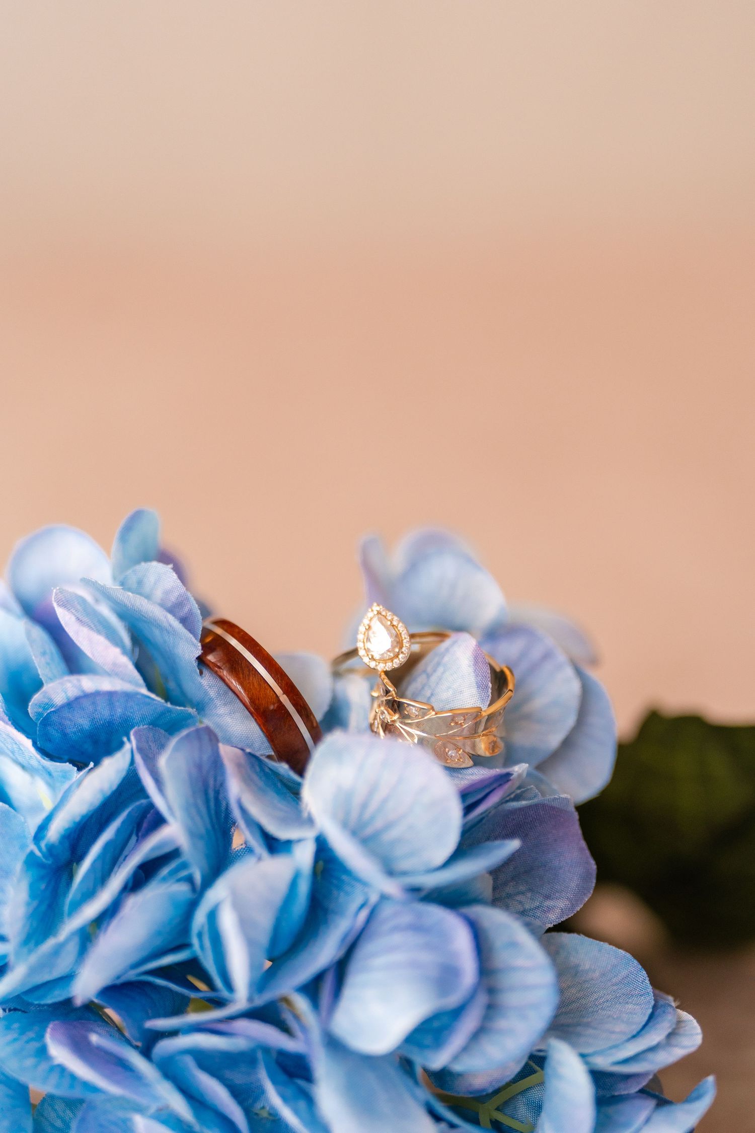 Gold pear-shaped diamond engagement ring and wooden wedding band resting on blue hydrangea flowers.