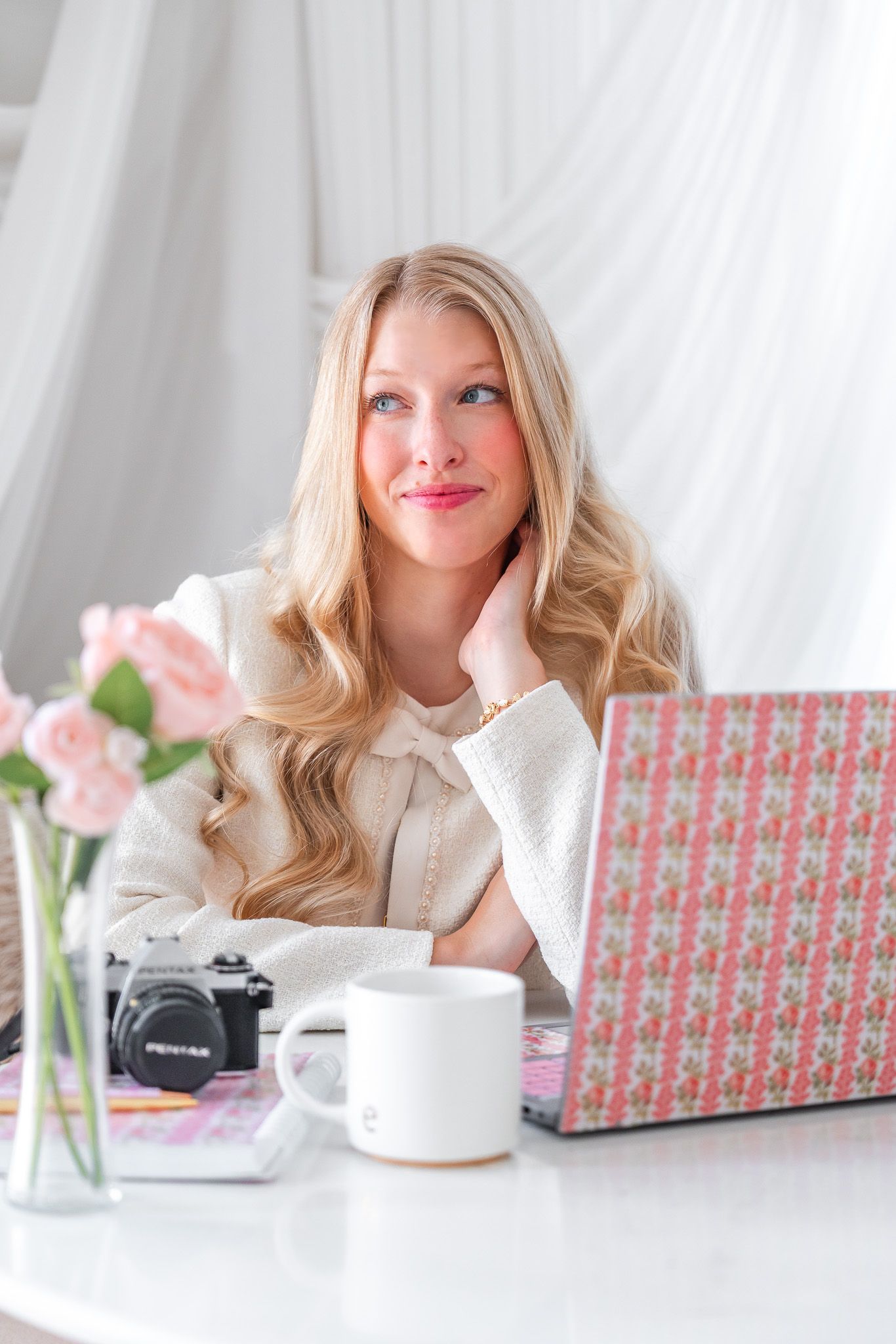 Blonde woman in white jacket sits at desk with laptop, roses, coffee mug, and vintage camera, gazing thoughtfully upward.