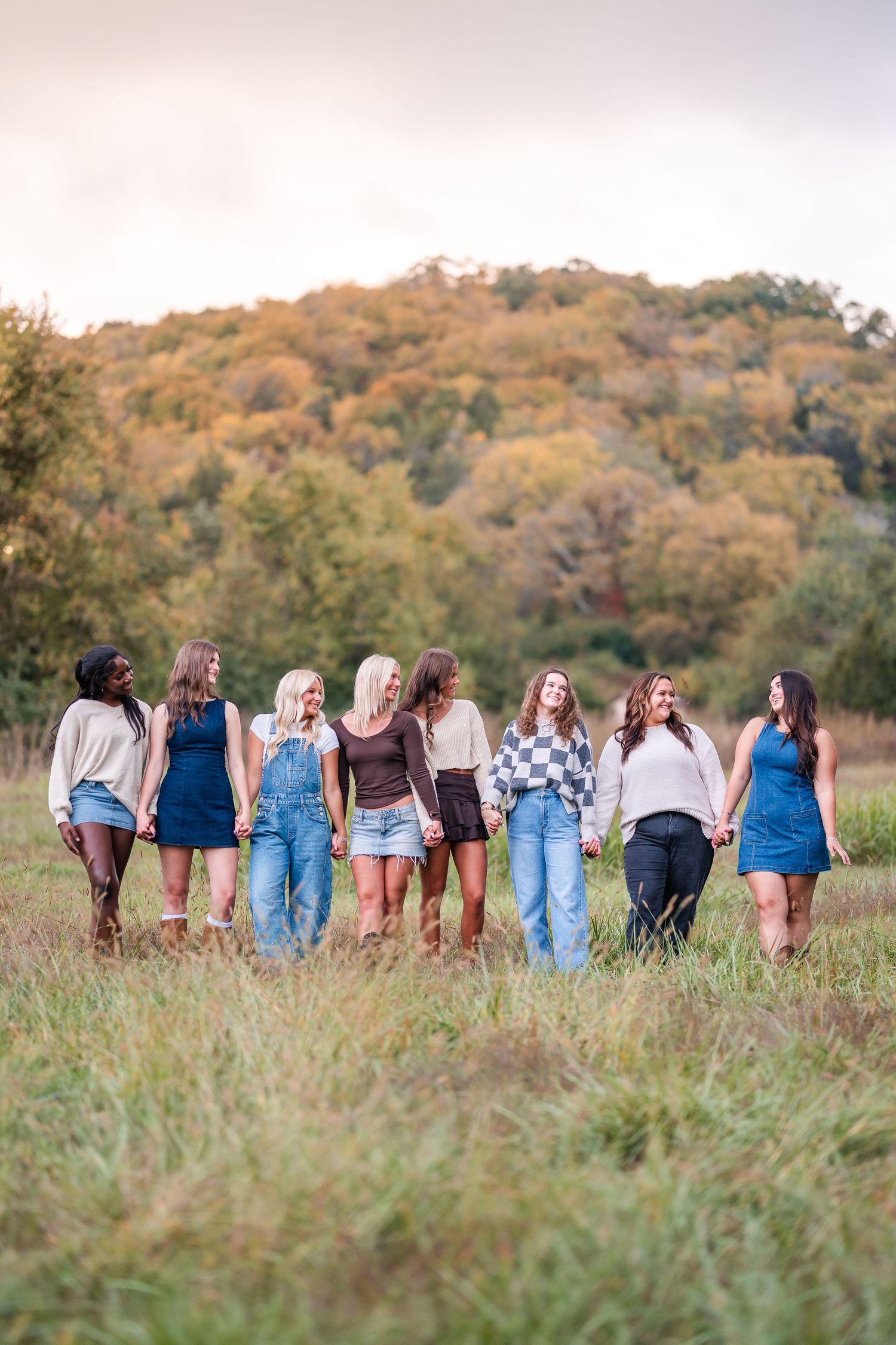 Group of eight young women holding hands walking through a field with autumn foliage backdrop.