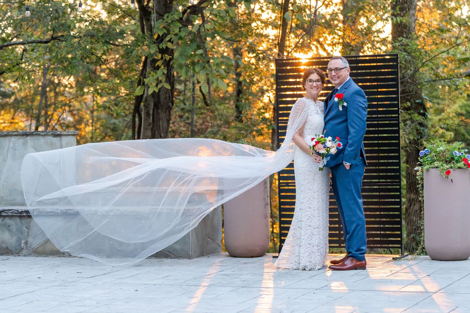Wedding couple embracing at sunset with bride's veil flowing dramatically in the breeze on stone patio.
