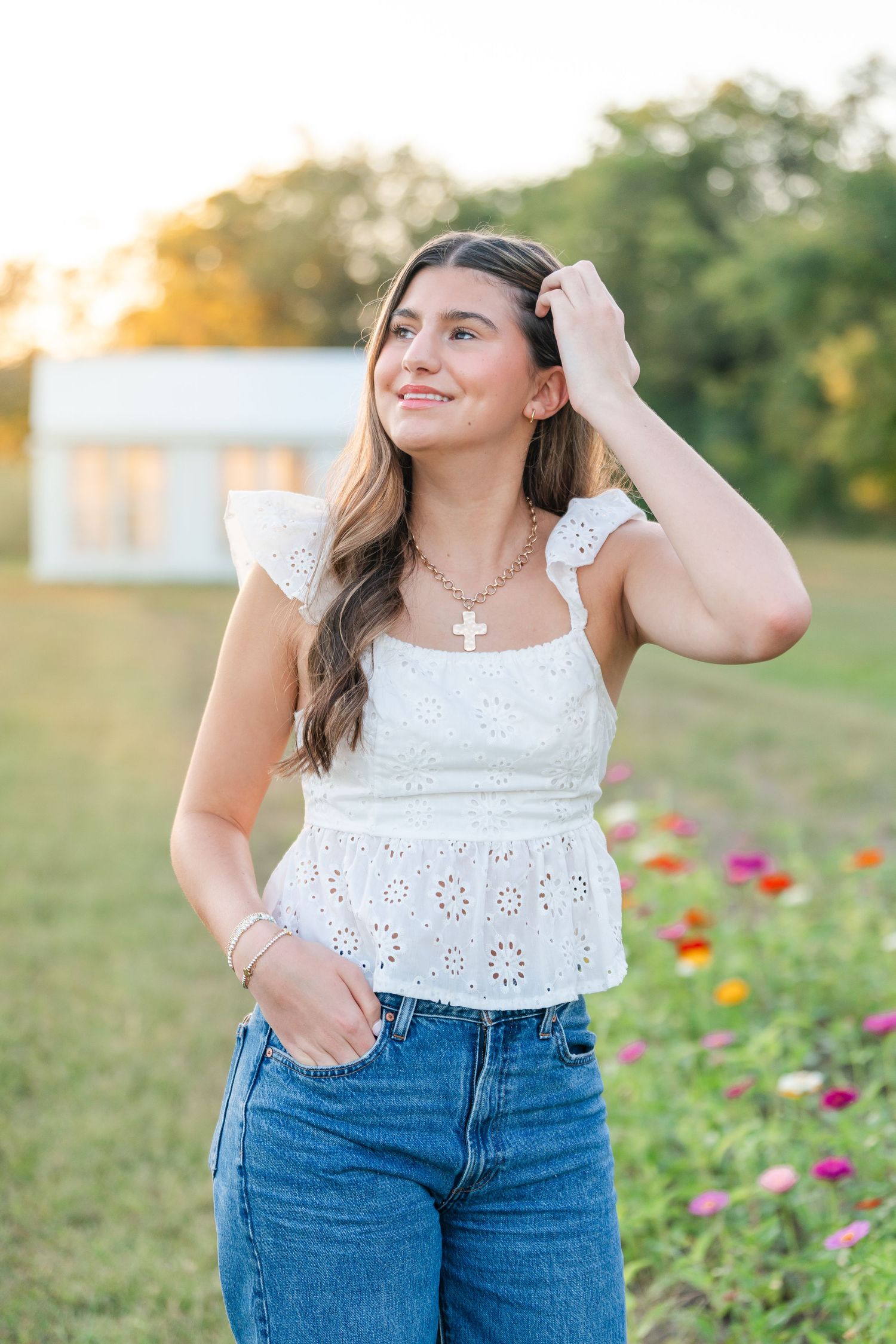 Smiling young woman in white eyelet top and jeans posing near flower garden at golden hour.