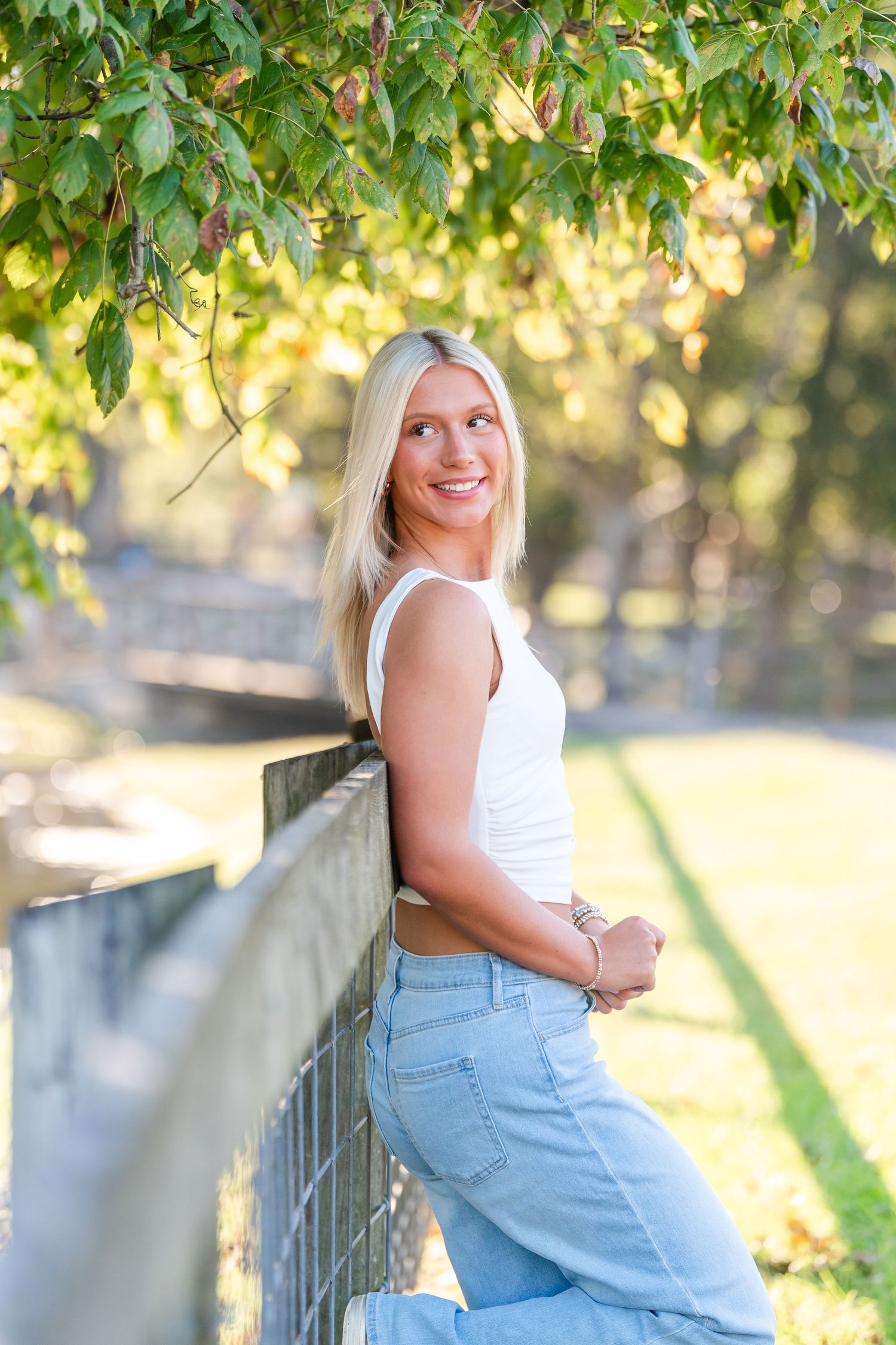 Blonde young woman in white tank top leaning on fence under green tree branches outdoors.