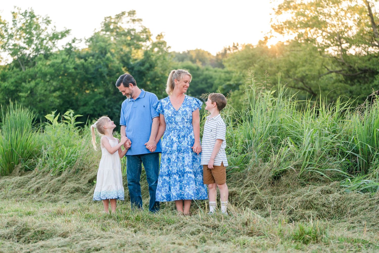 Family of four standing together in golden sunset light beside tall green grass in open field.
