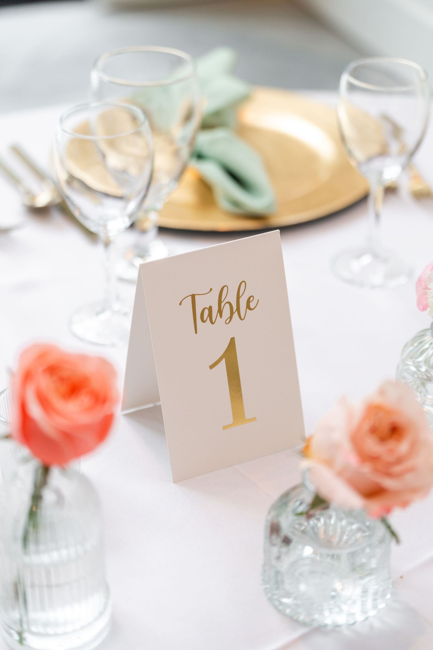 Gold-script Table 1 card displayed at a wedding reception table with coral roses, gold chargers, and crystal glassware.