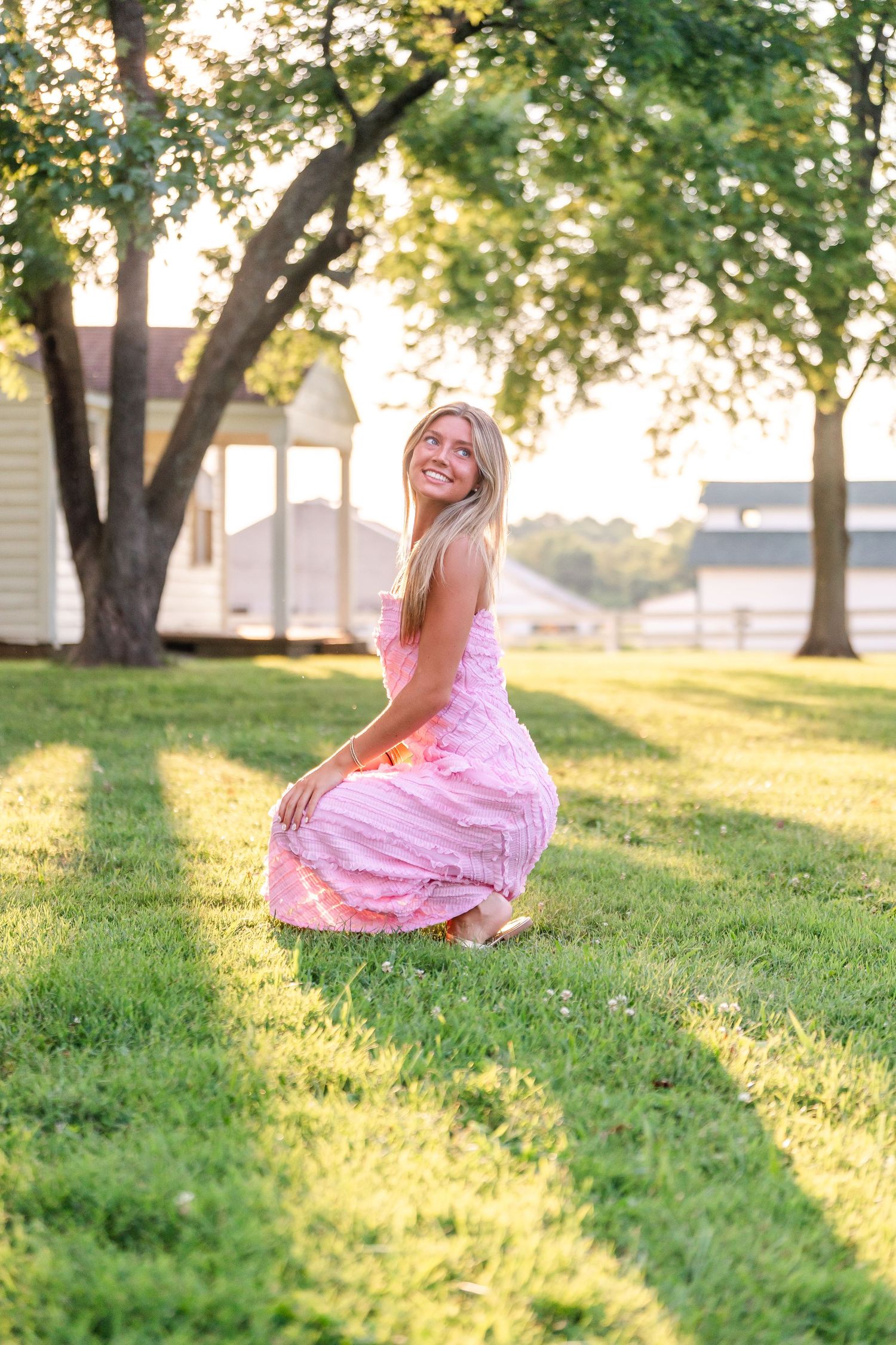 Smiling blonde woman in pink gingham maxi dress crouching on green grass in golden afternoon sunlight.