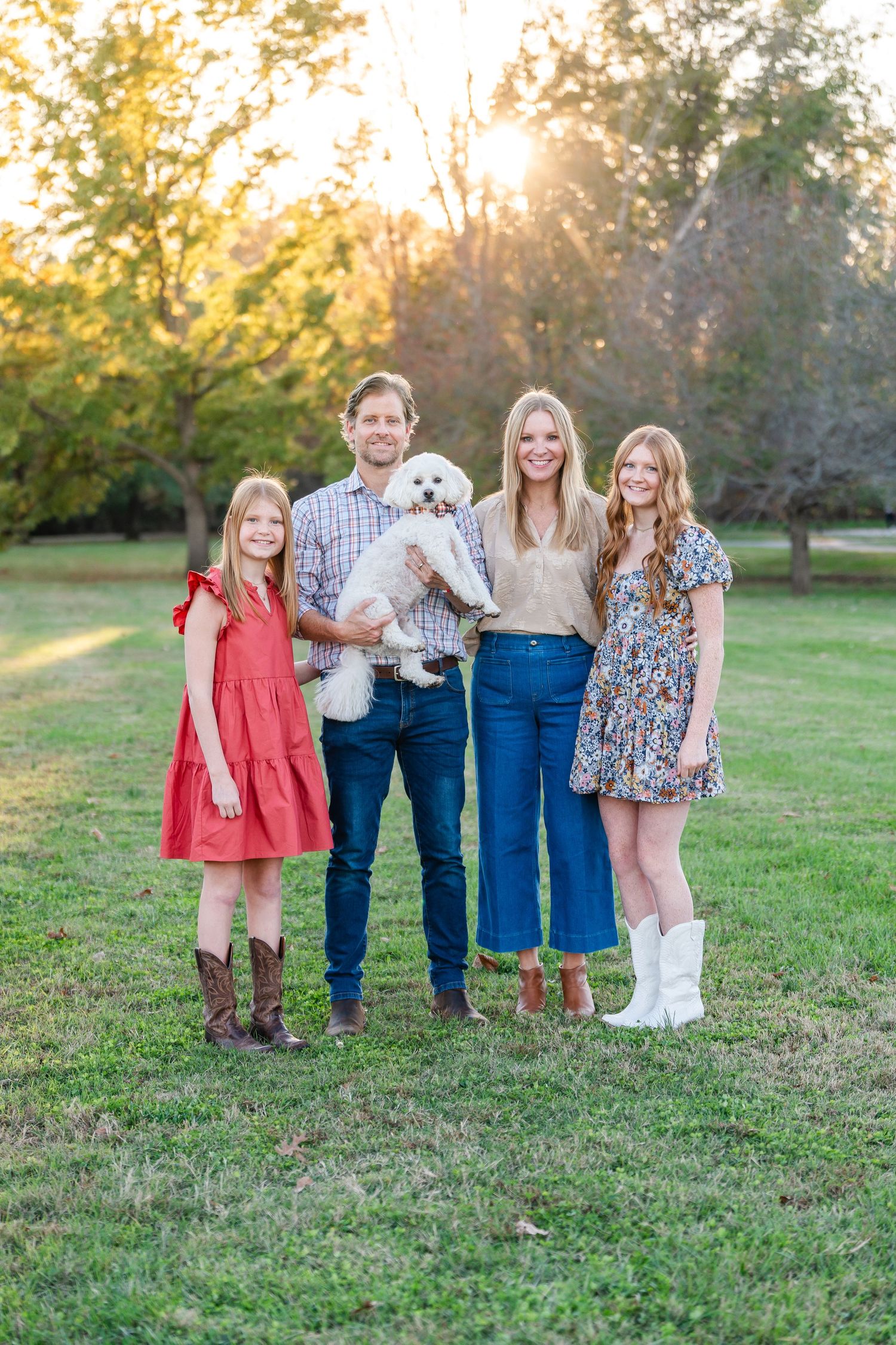 Family of four posing with a white fluffy dog in a sunlit park during golden hour.