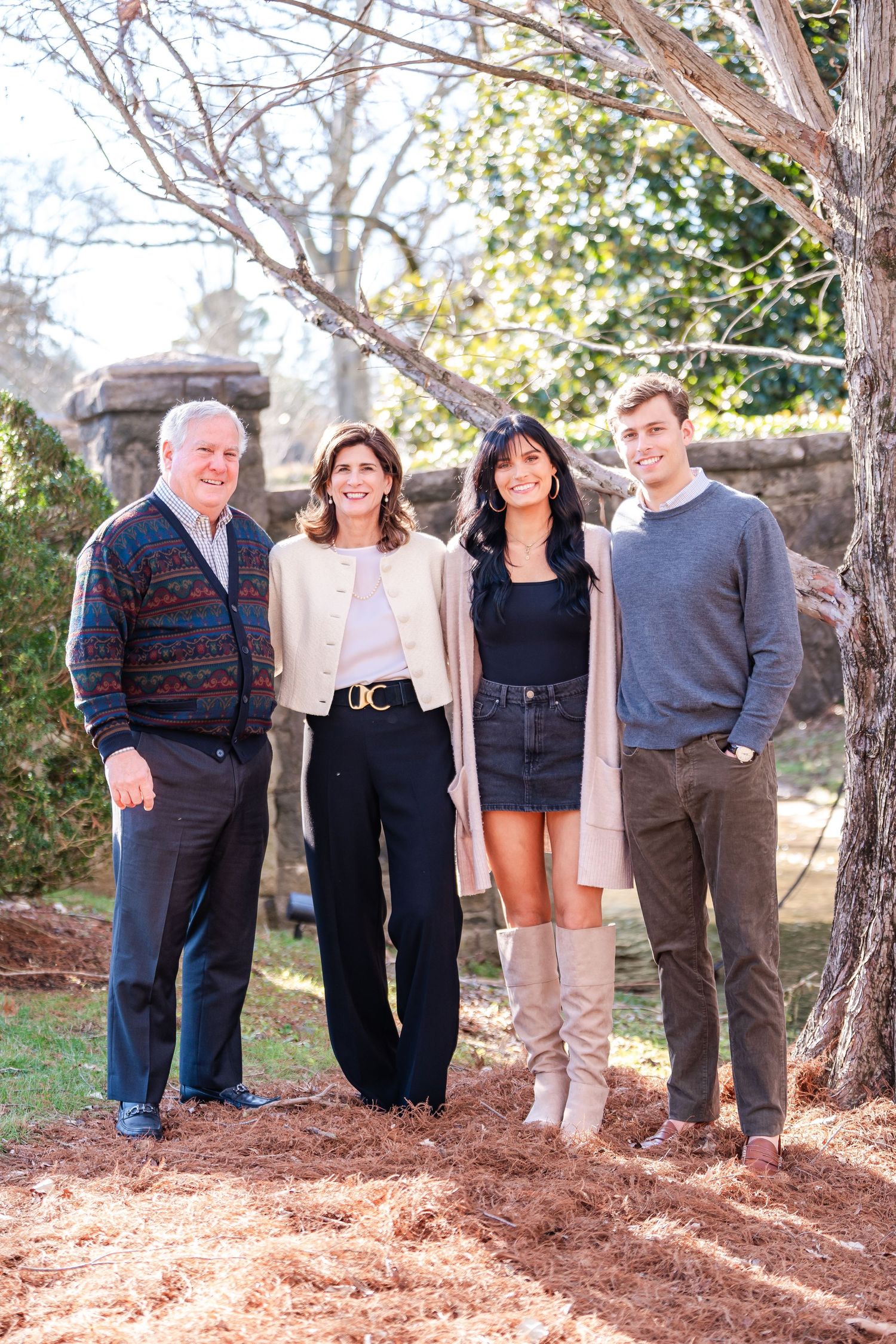 Four smiling family members pose together outdoors near trees and rocks on a sunny day.