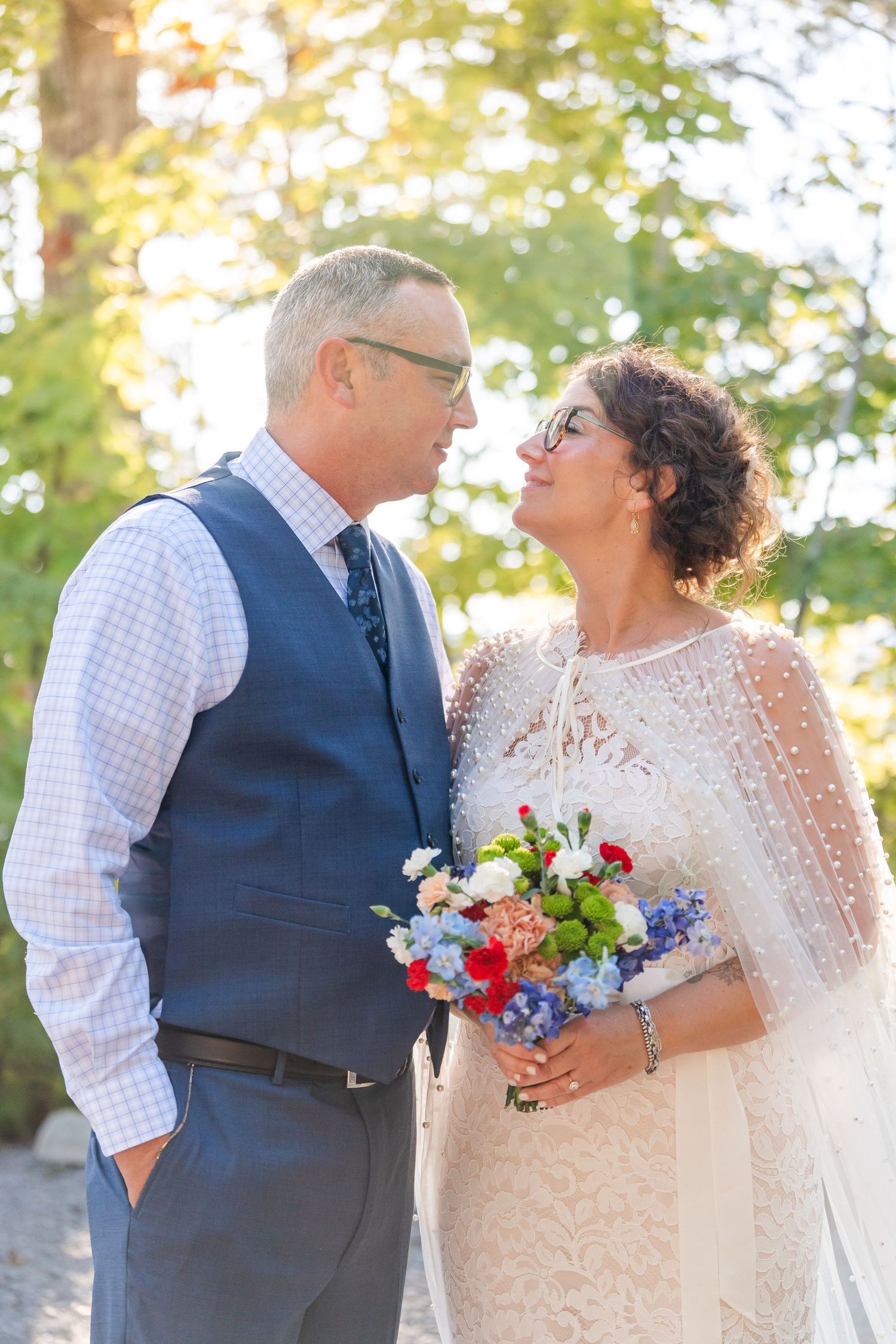 Older couple sharing a tender moment outdoors, bride in lace cape gown holding red and blue bouquet.