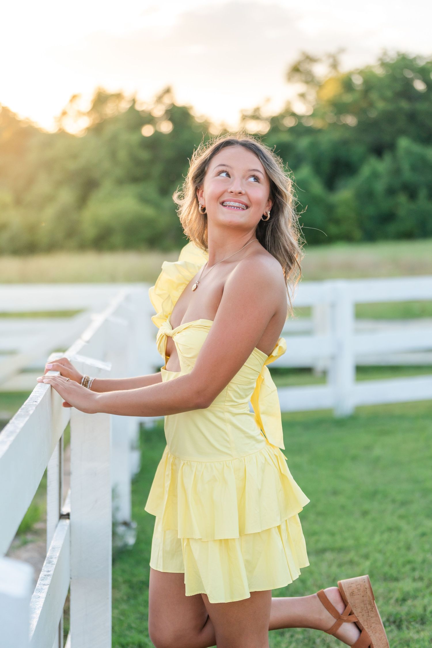 Smiling young woman in yellow ruffled dress leaning on white fence during golden hour sunset.