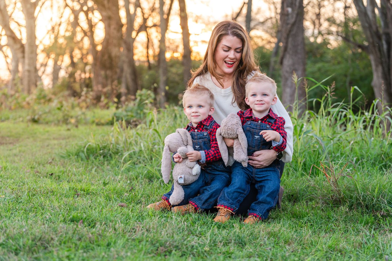 Mother smiling and hugging her twin toddler boys holding stuffed animals in a sunny park.