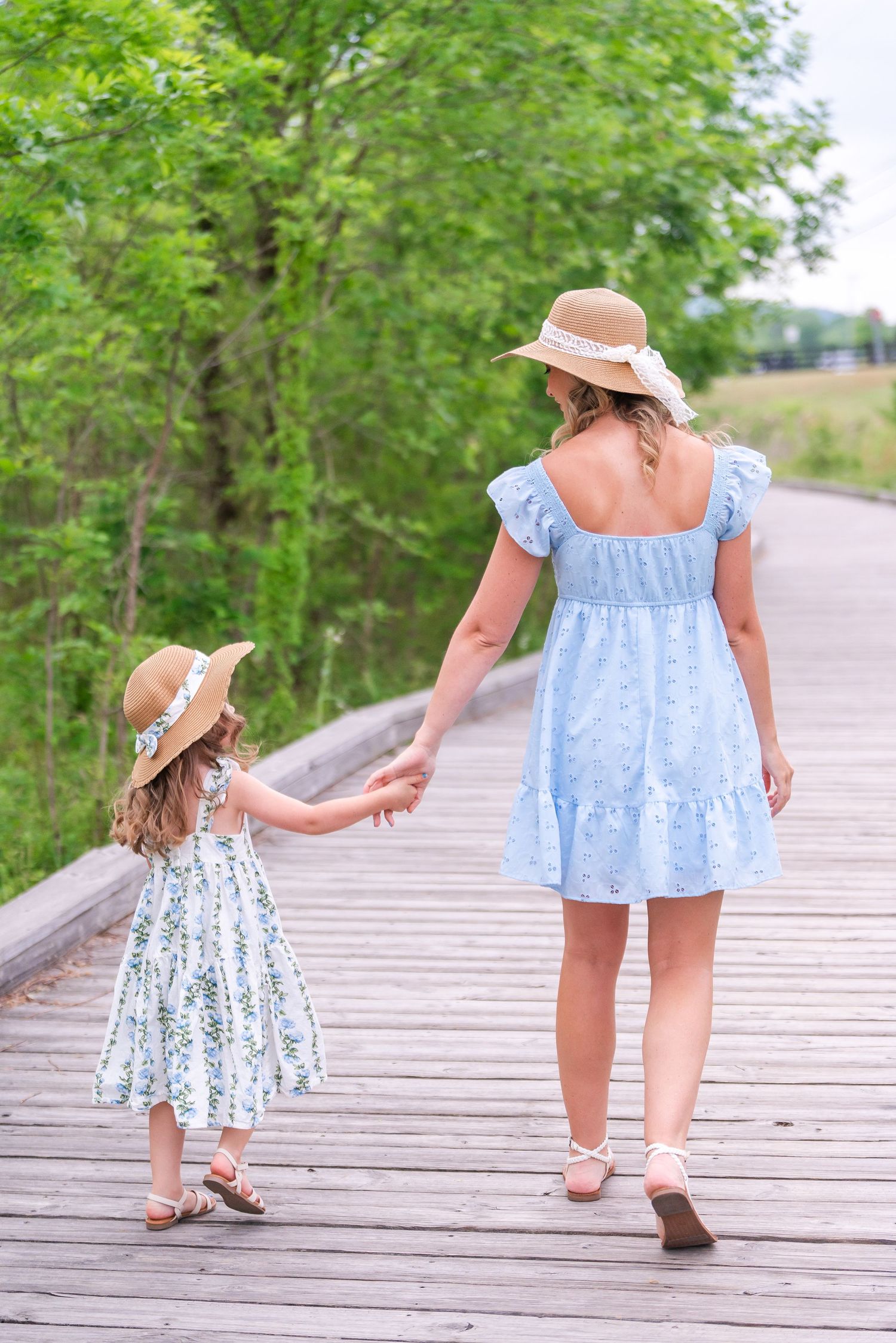 Mother and daughter in matching blue floral dresses and straw hats walking hand in hand on a wooden boardwalk.