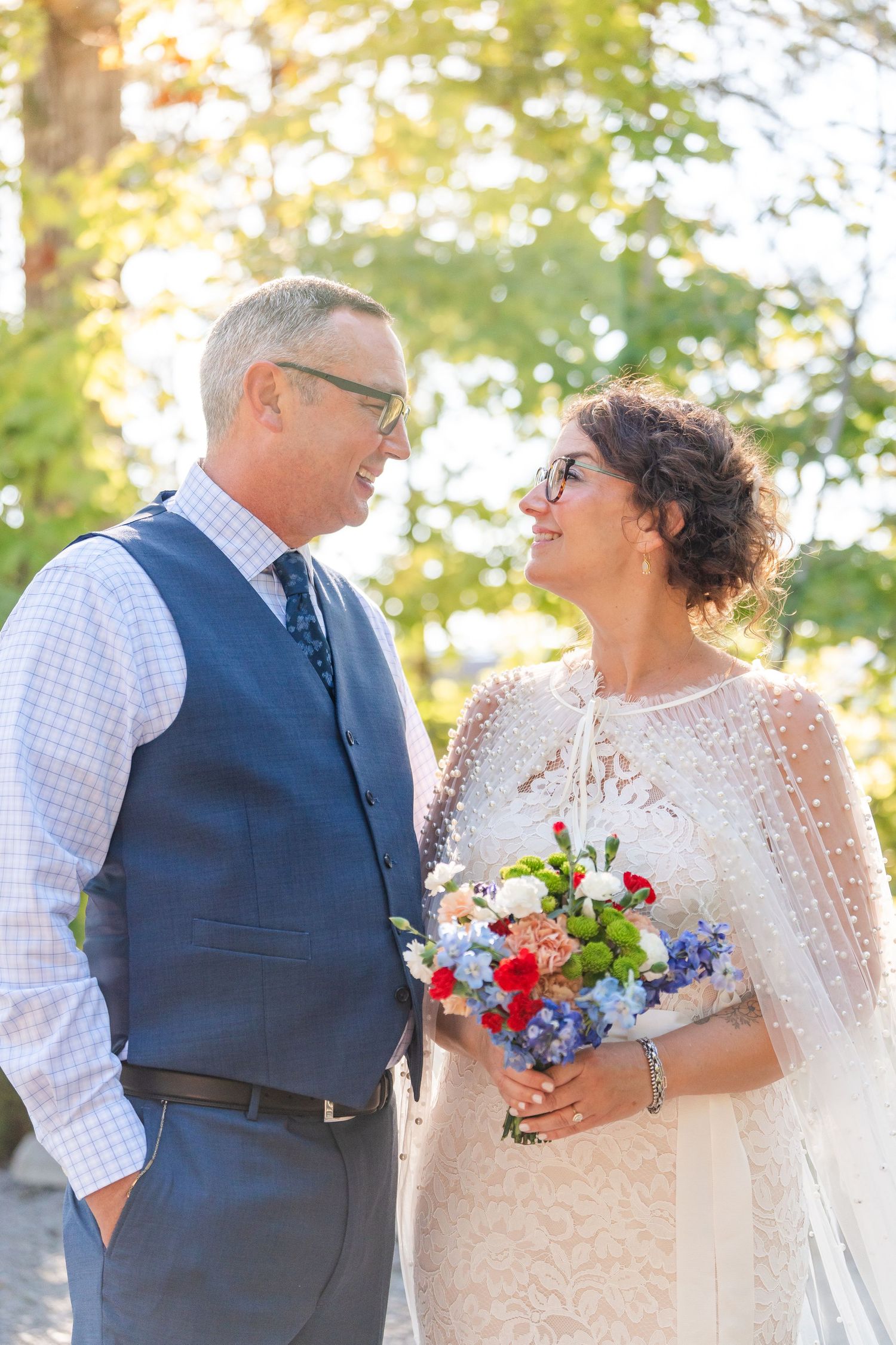 Mature bride and groom gazing at each other outdoors, bride holding colorful bouquet, groom in blue vest.