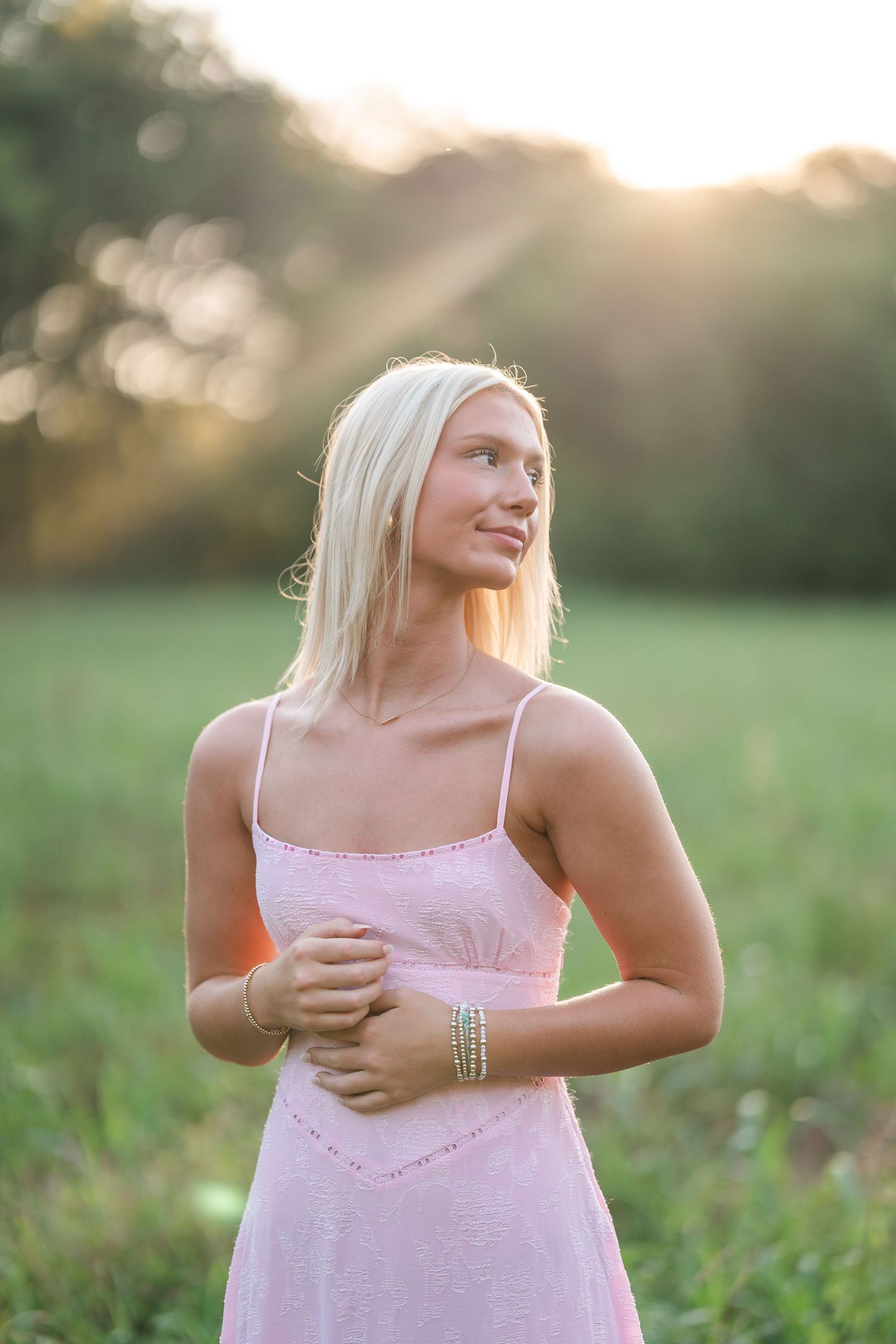 Blonde young woman in pink sundress standing in green field with warm sunset rays behind her.