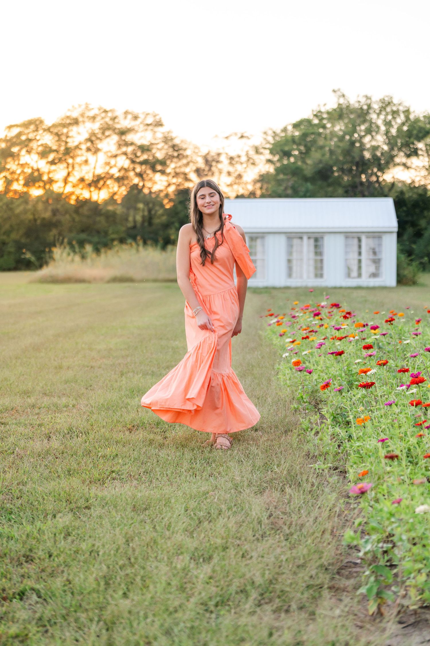 Young woman in flowing peach gown walking beside colorful flower garden at golden hour with white barn in background.