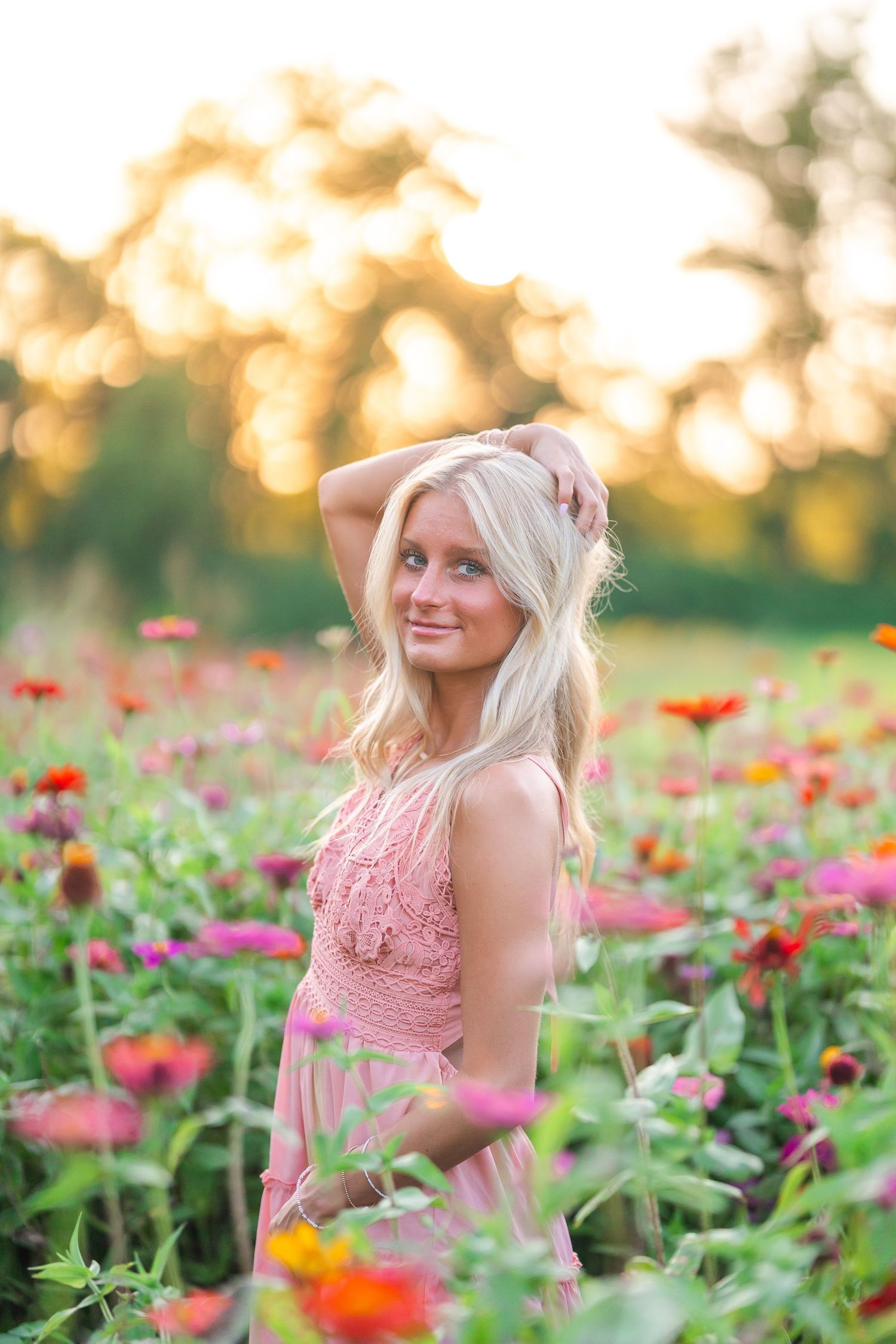 Blonde woman in pink dress posing in colorful zinnia flower field at golden sunset.