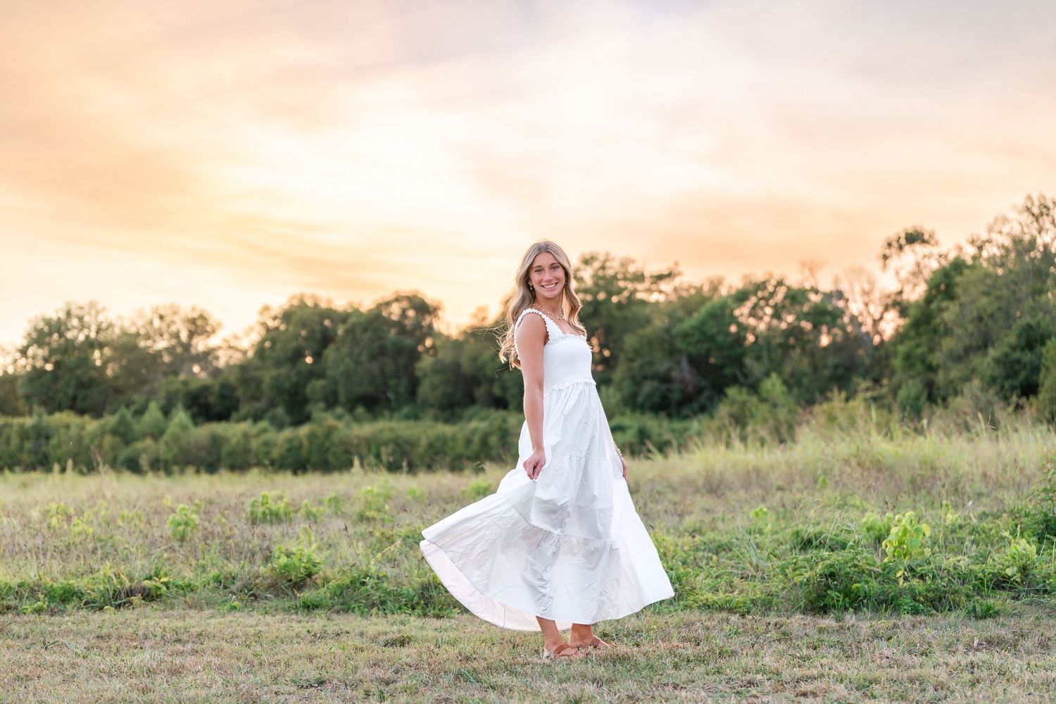 Woman in flowing white maxi dress spinning in open field during dramatic golden sunset sky.