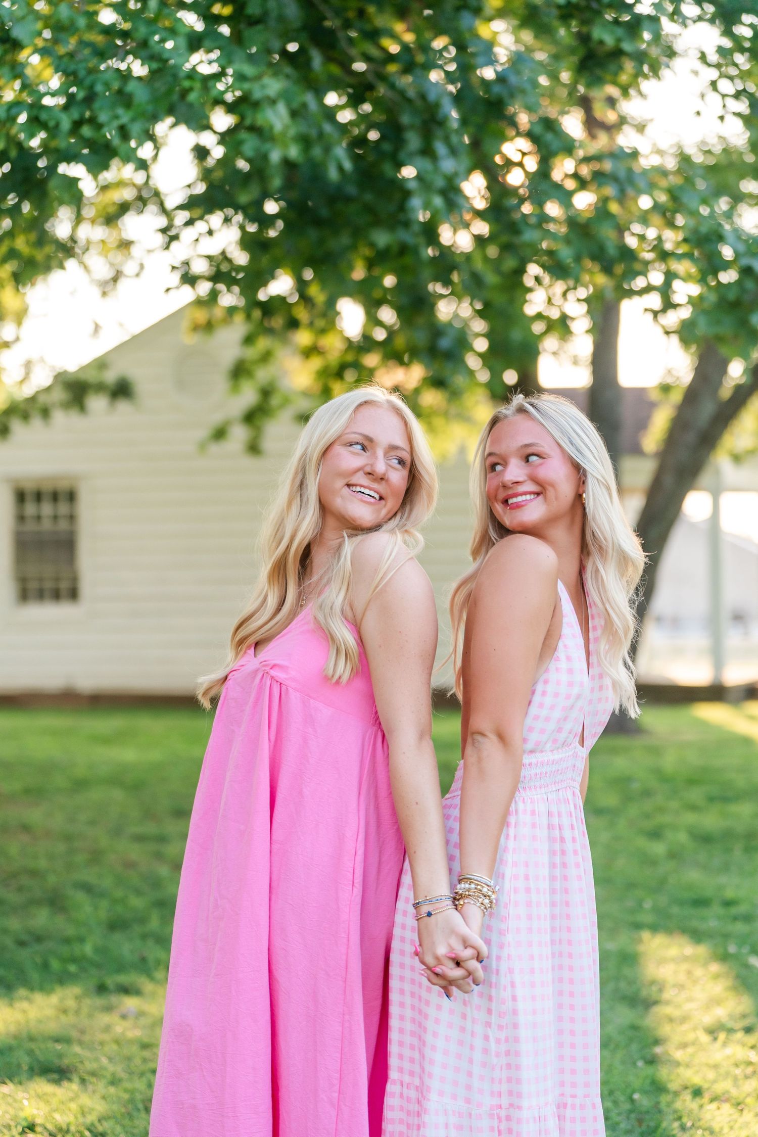 Two smiling blonde women in pink dresses holding hands and posing in sunny green park.
