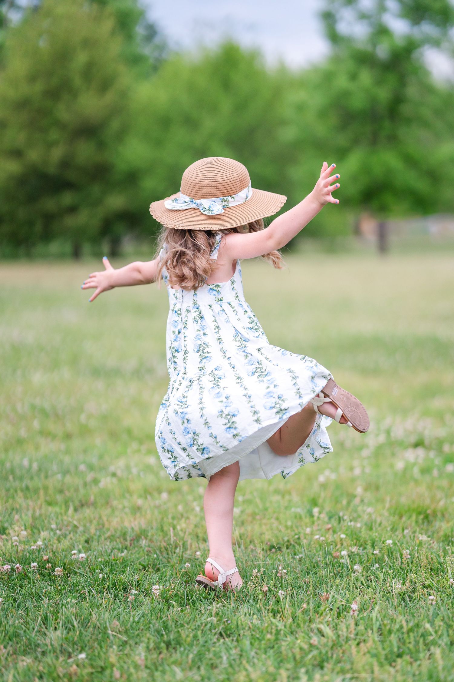 Young girl in floral dress and straw hat twirling joyfully in a green meadow on a sunny day.