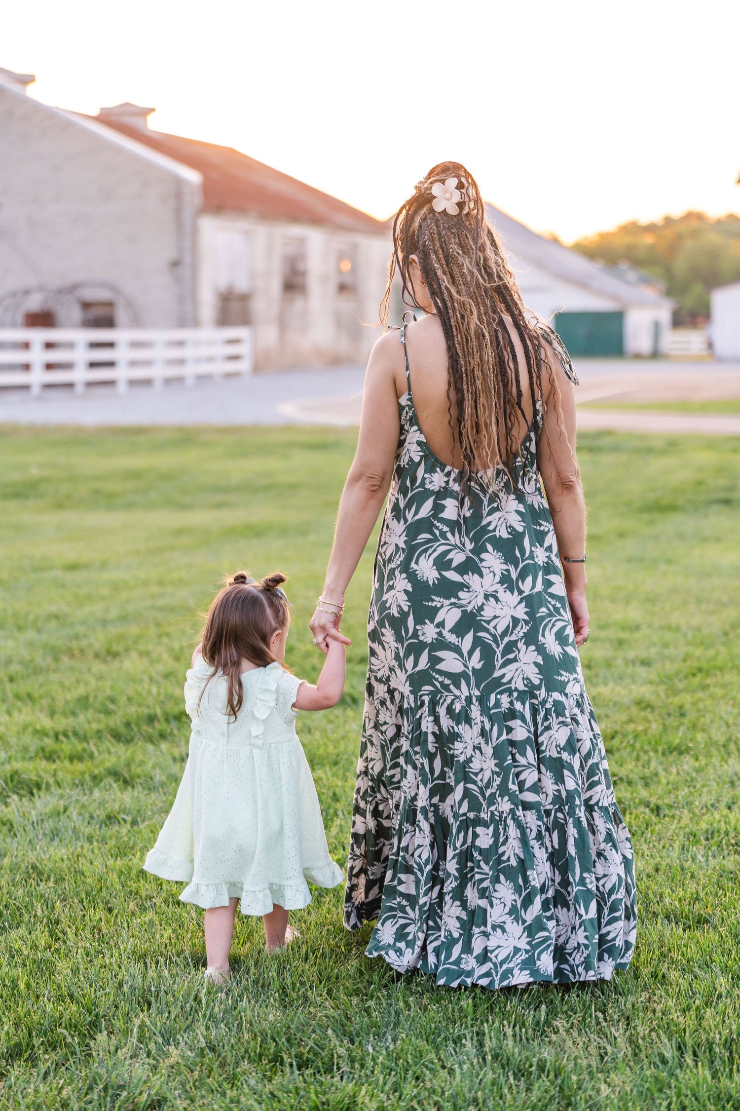 Mother with dreadlocks in floral maxi dress holds hands with toddler girl in white dress walking on green grass.