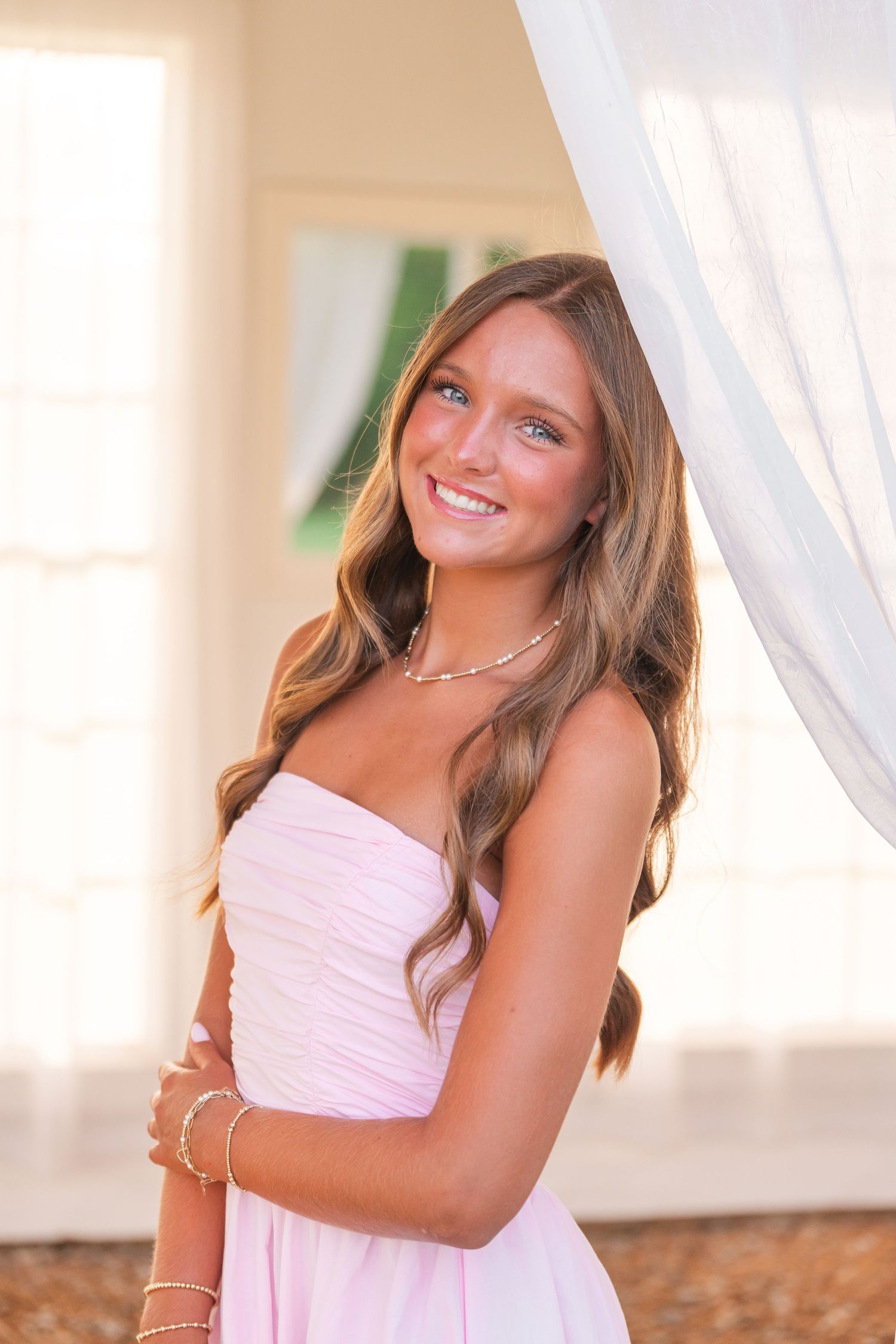 Young woman in pink strapless dress with long wavy hair, smiling brightly near sheer white curtains.