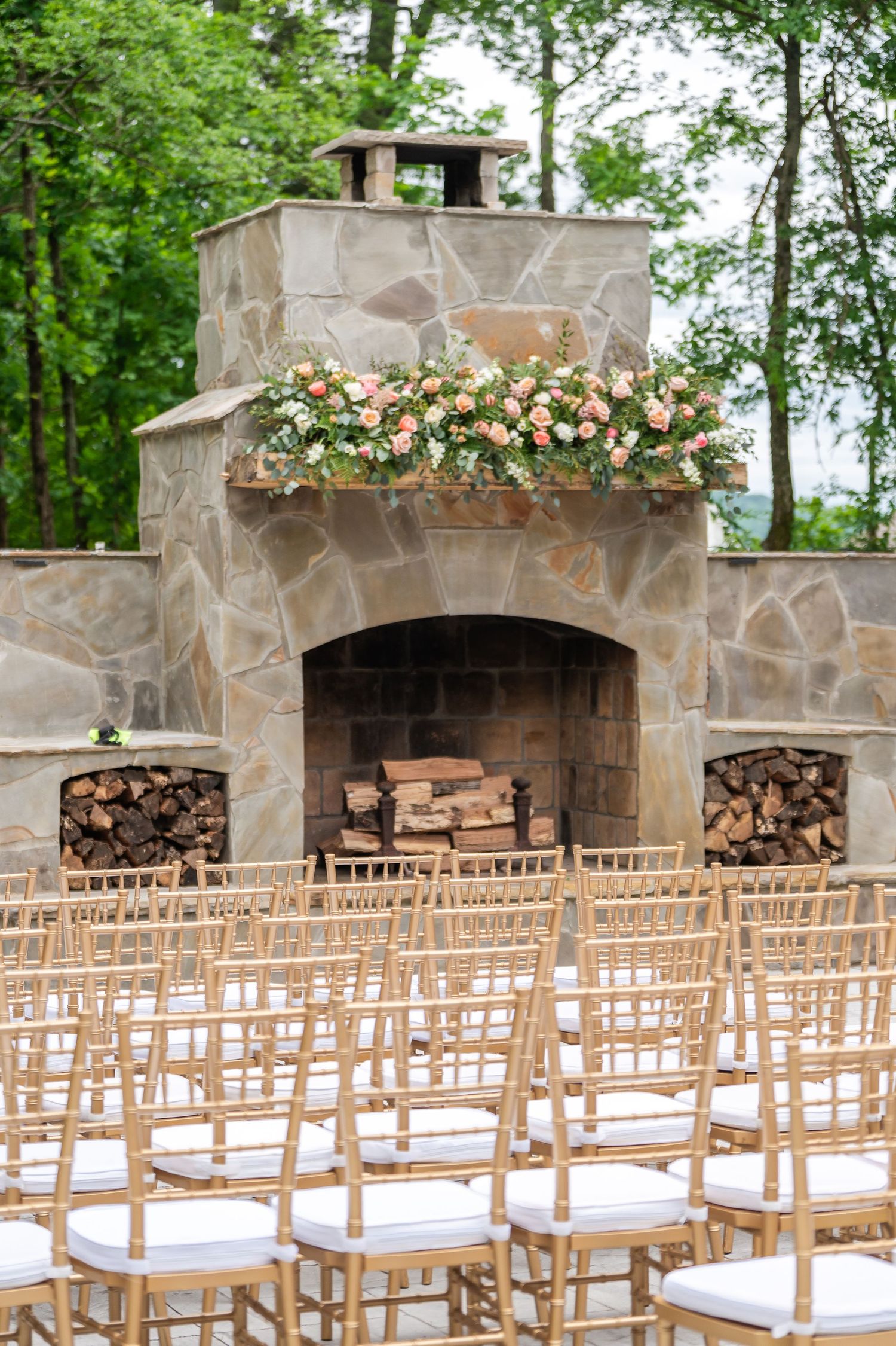 Elegant outdoor wedding ceremony setup with gold chiavari chairs facing a stone fireplace adorned with pink floral arrangements.