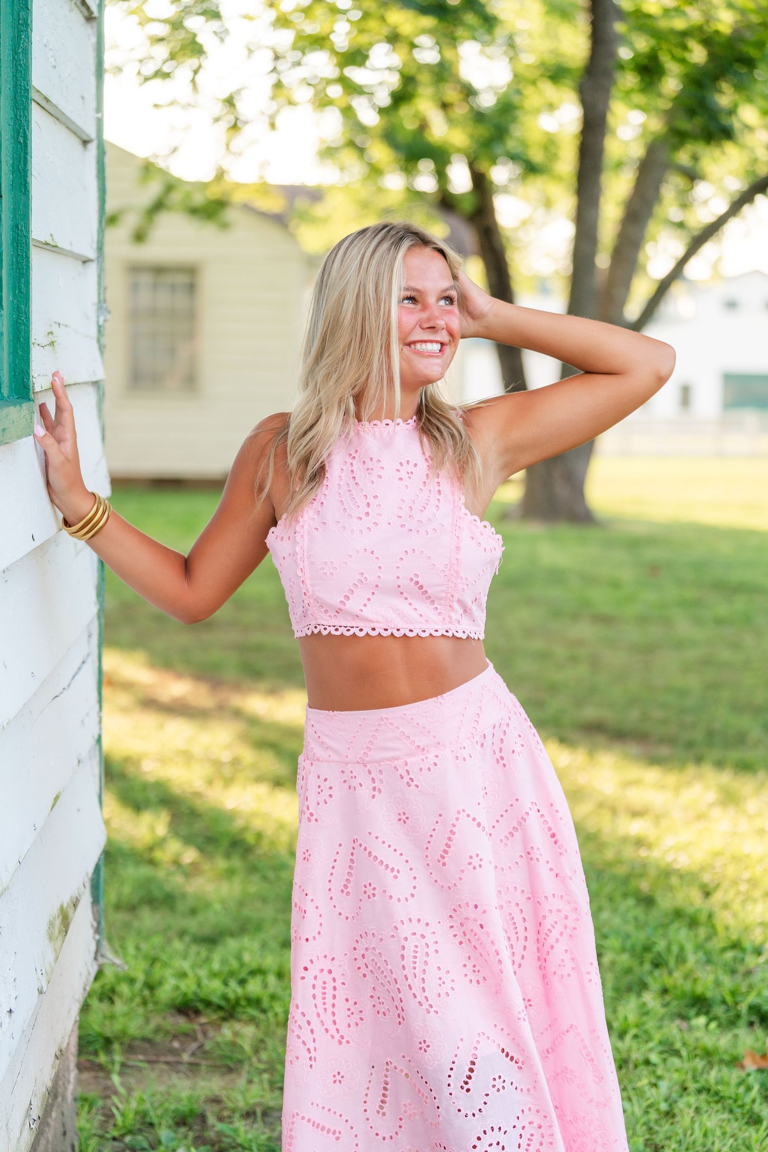 Smiling blonde woman in pink crop top and skirt posing against white building outdoors.