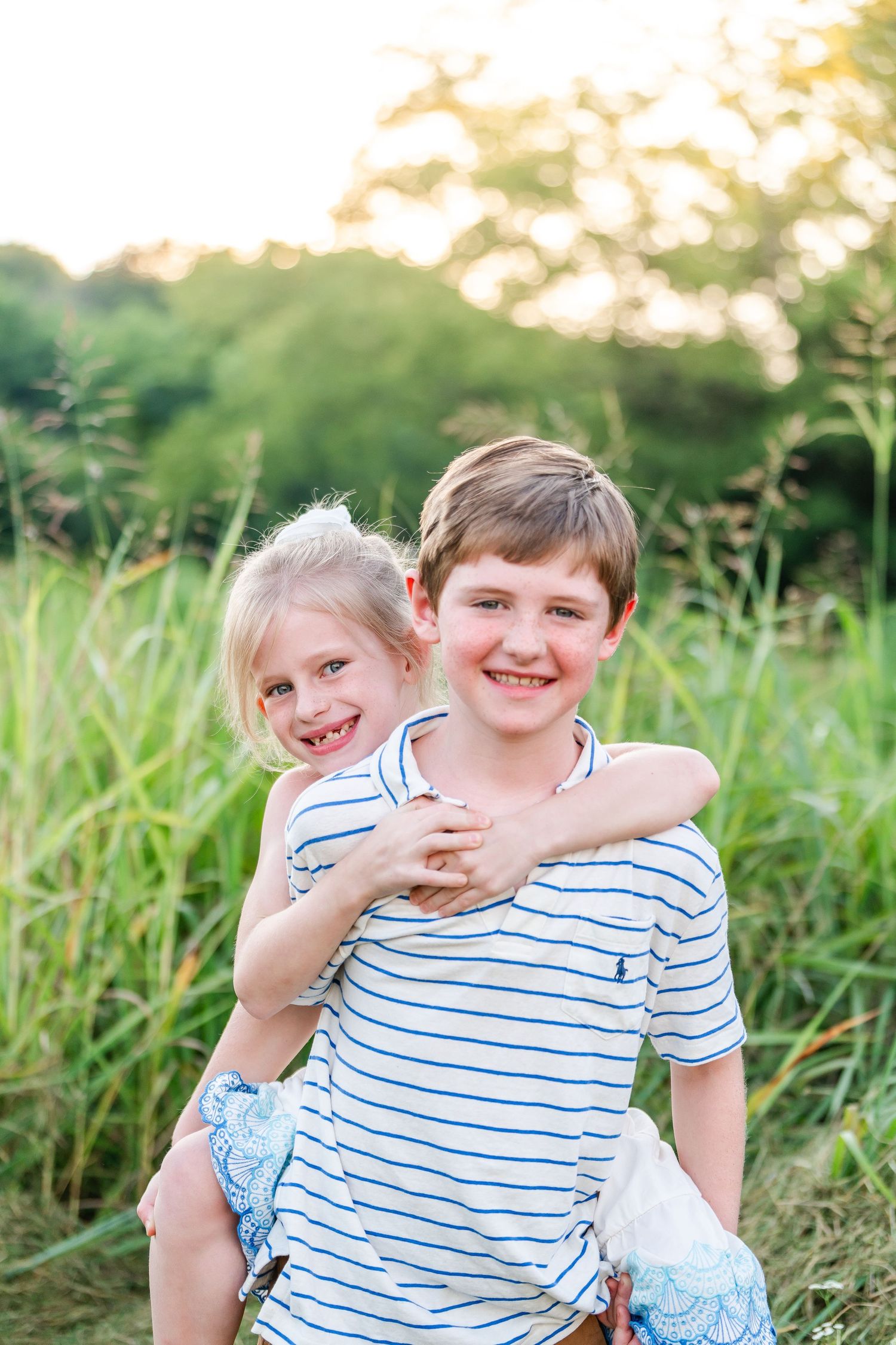 Smiling boy holding his little sister piggyback in a sunny green outdoor setting.