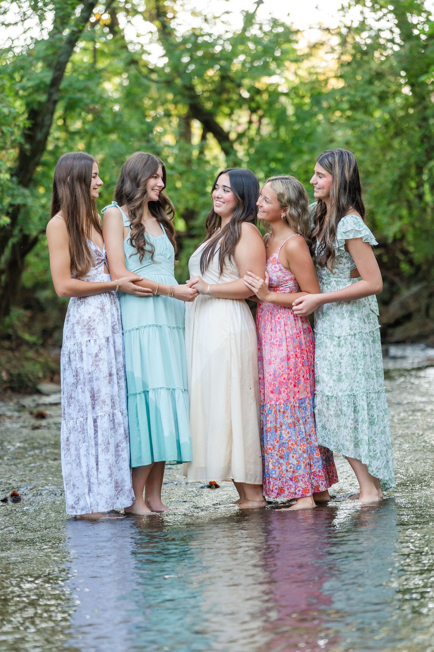 Five young women in colorful floral maxi dresses standing together in a shallow creek surrounded by trees.