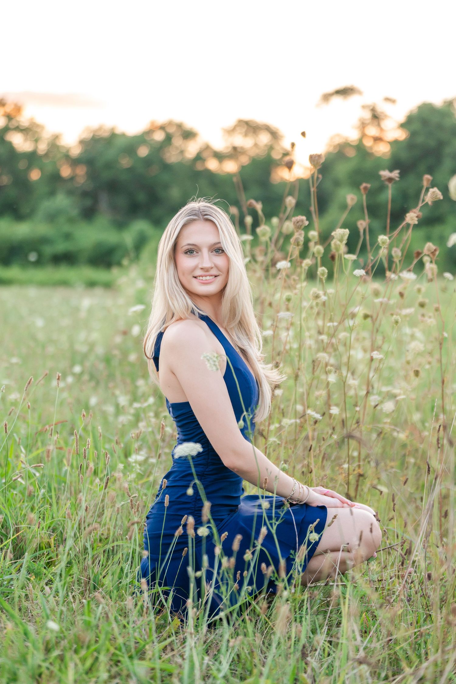 Blonde teen girl in navy dress sitting elegantly in a sunlit wildflower field at golden hour.