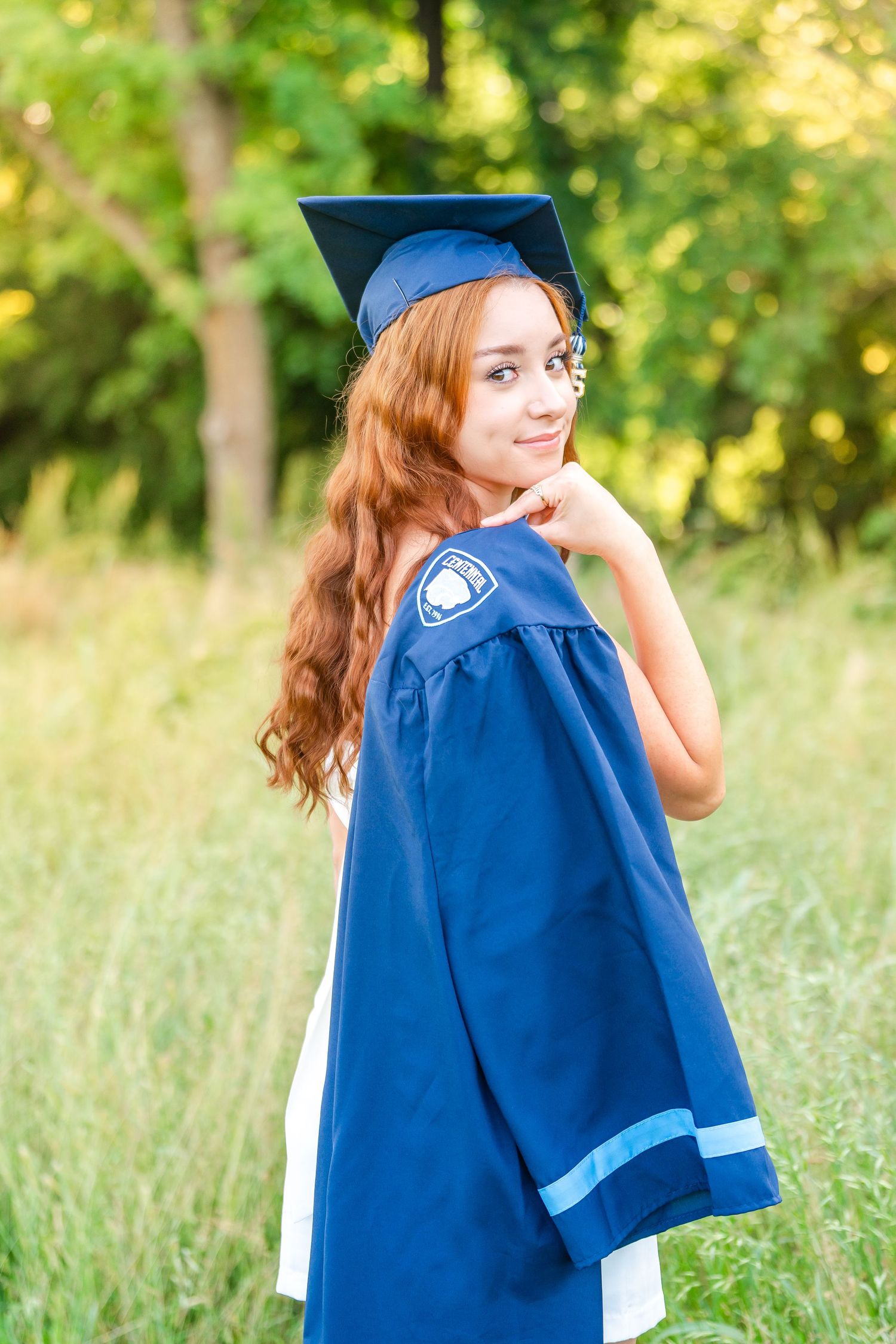Smiling redhead girl in blue graduation cap and gown posing outdoors in green field.