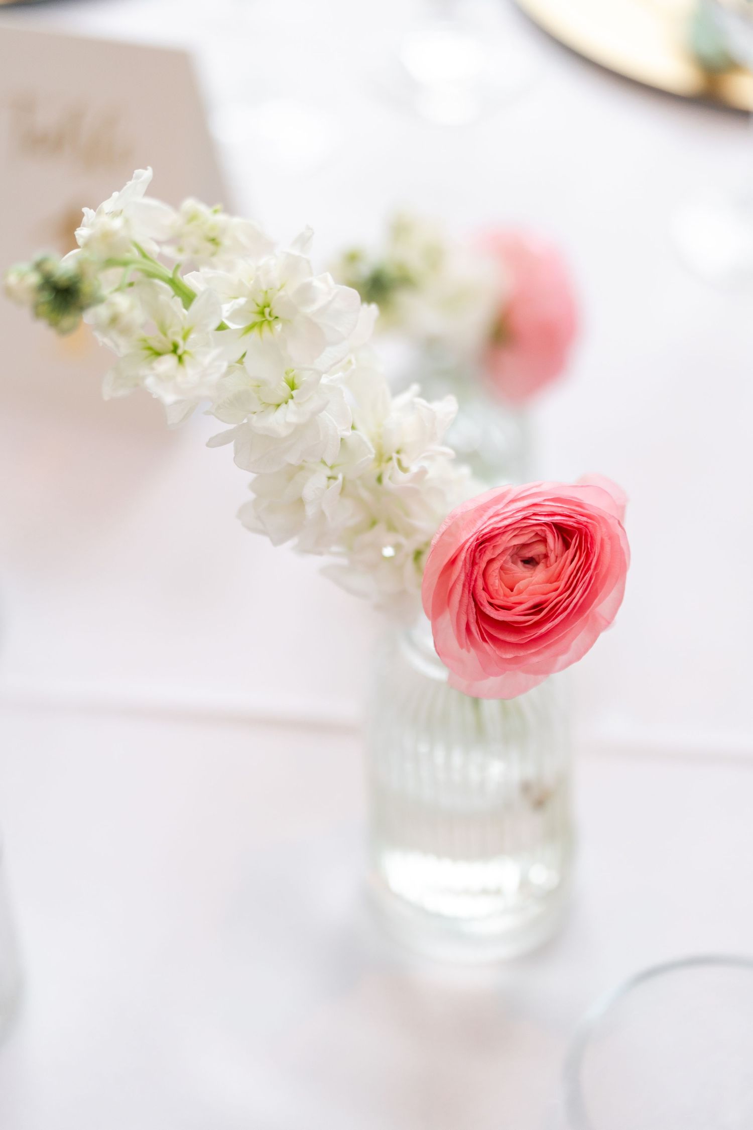 Elegant wedding table centerpiece with pink ranunculus and white stock flowers in a small glass vase.