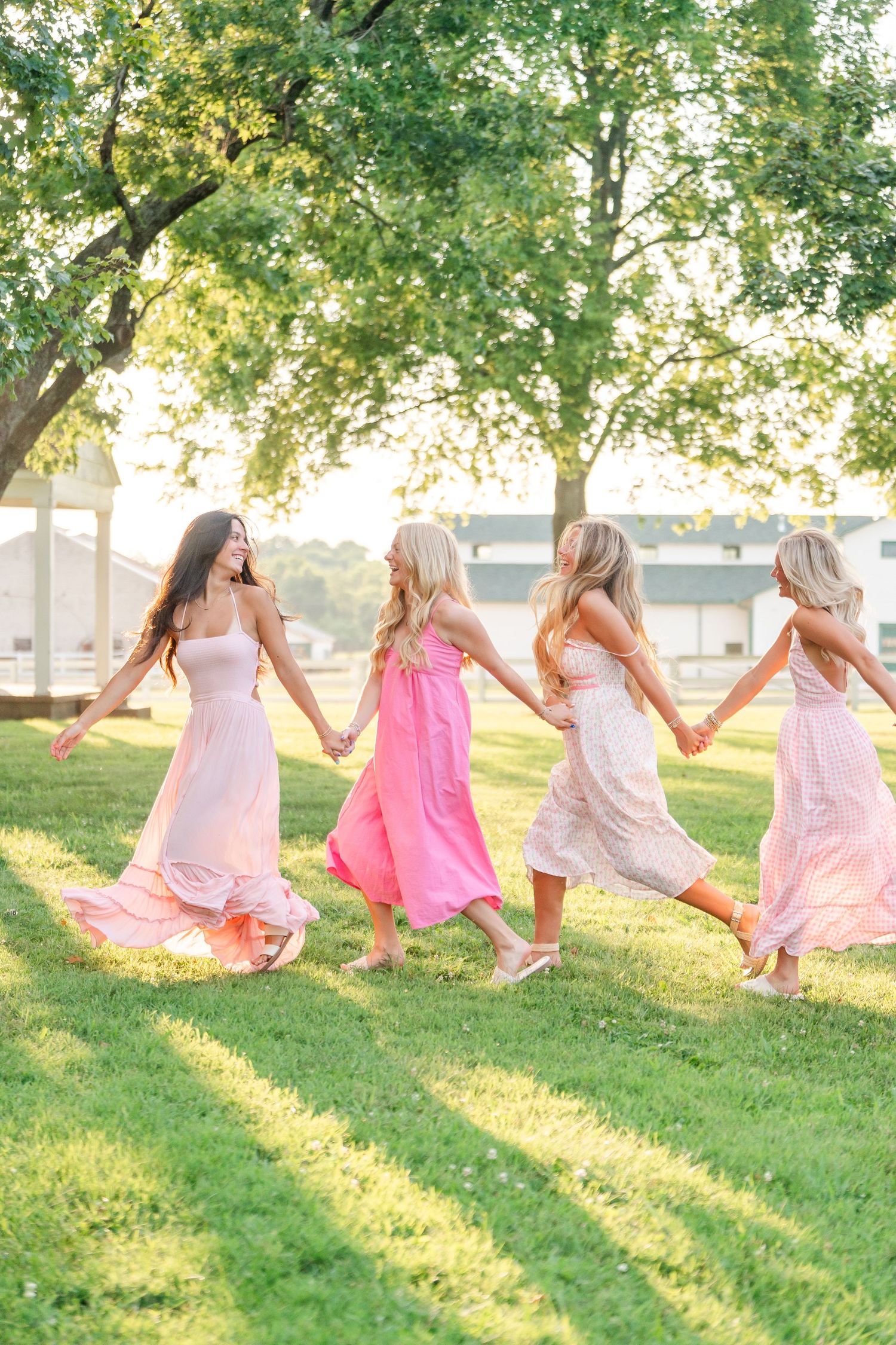 Four young women in pink dresses holding hands walking through green park at sunset.