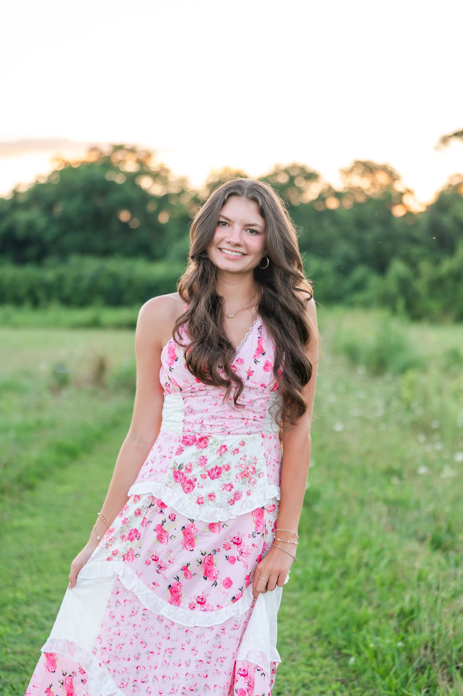 Young woman with long brown hair in pink floral halter dress smiling in green field at sunset.