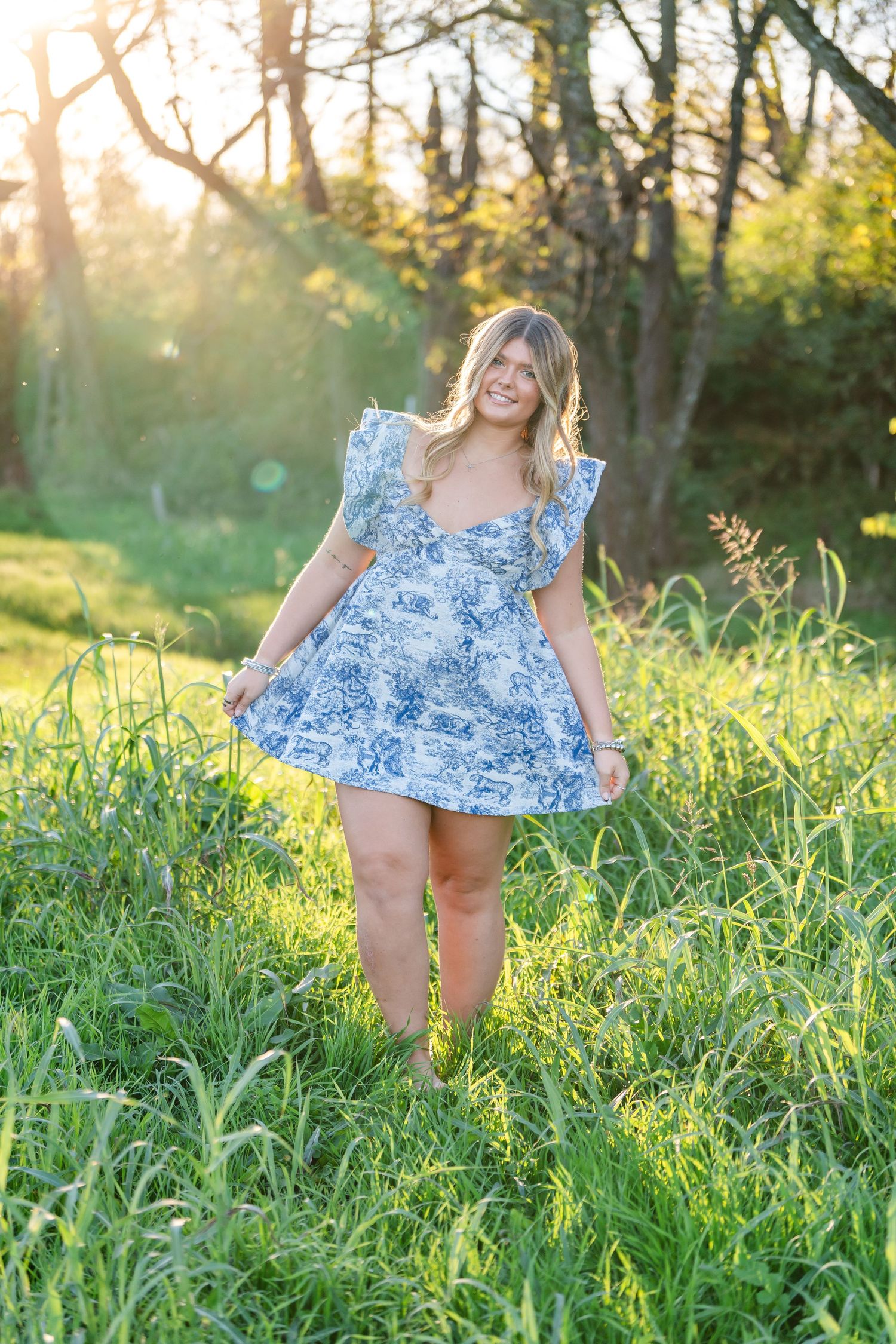 Young woman in blue floral dress twirling in green meadow during golden hour sunset photography session.