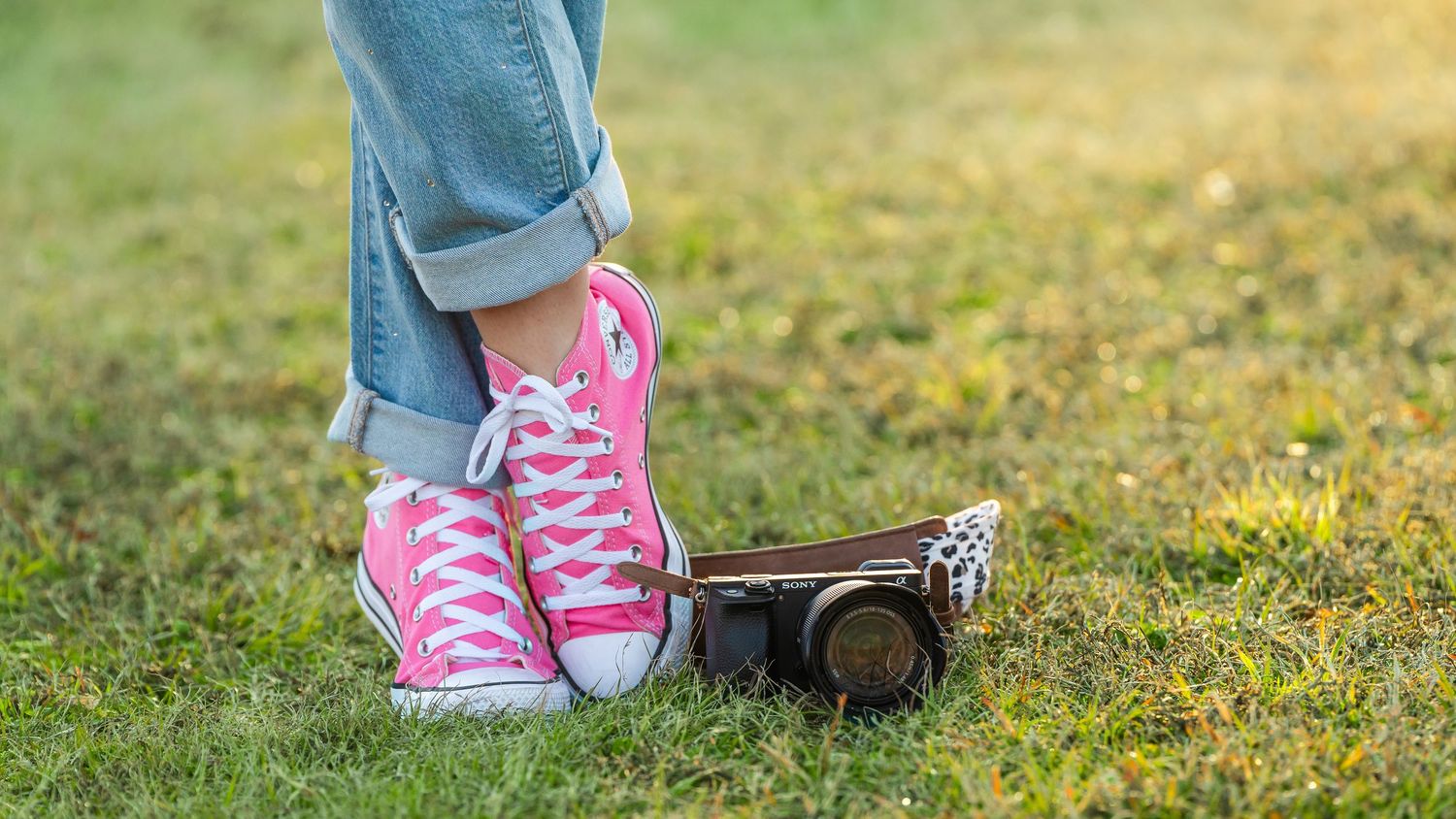 Pink Converse sneakers with rolled jeans next to a camera on green grass.