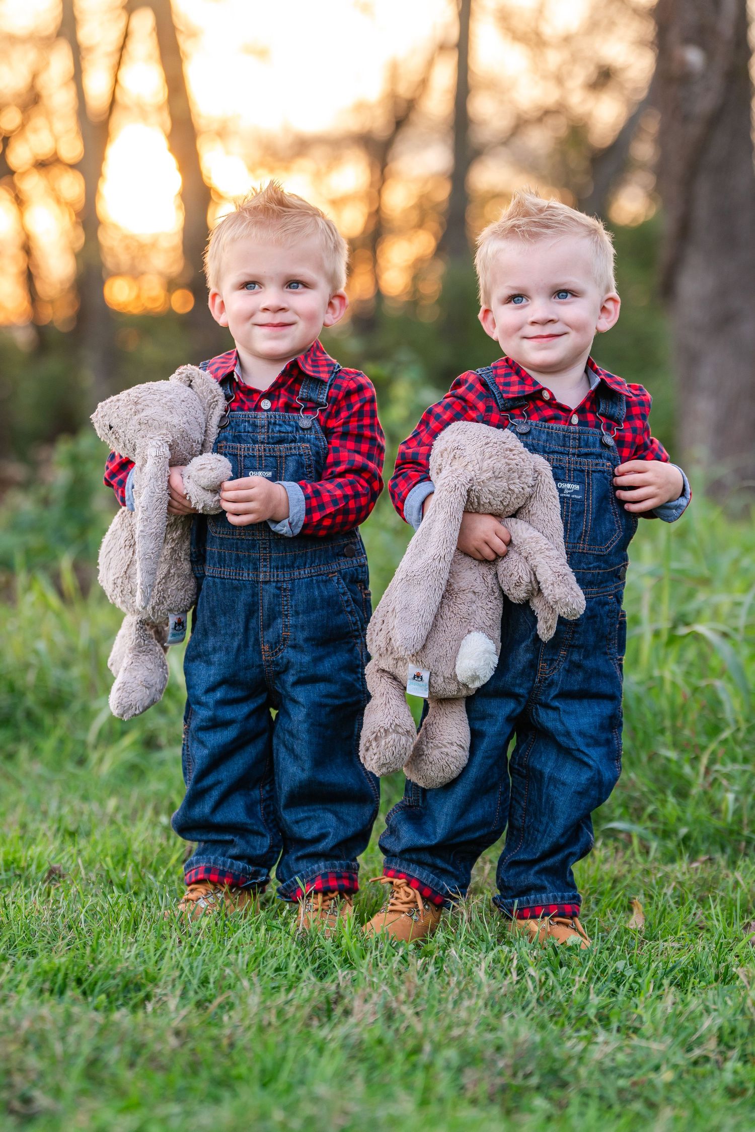 Twin toddler boys in matching red plaid shirts and denim overalls holding stuffed animal toys outdoors.