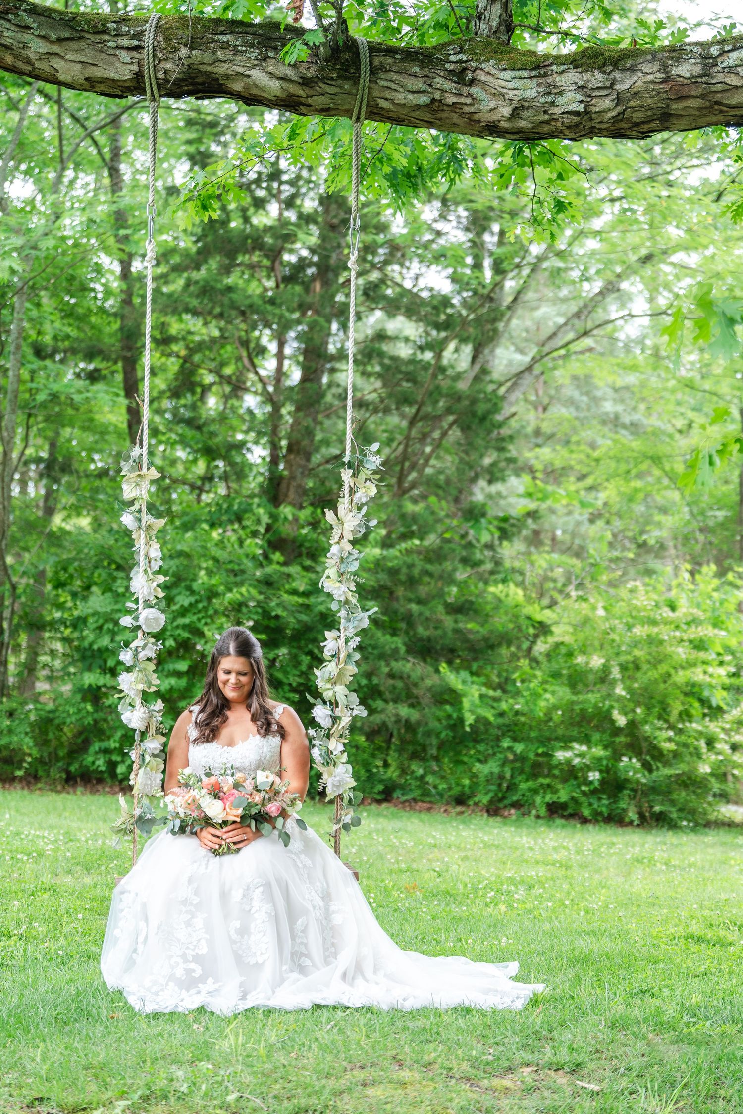 Bride in a lace ball gown sitting on a floral-decorated swing beneath a tree, holding a peach and white bouquet.