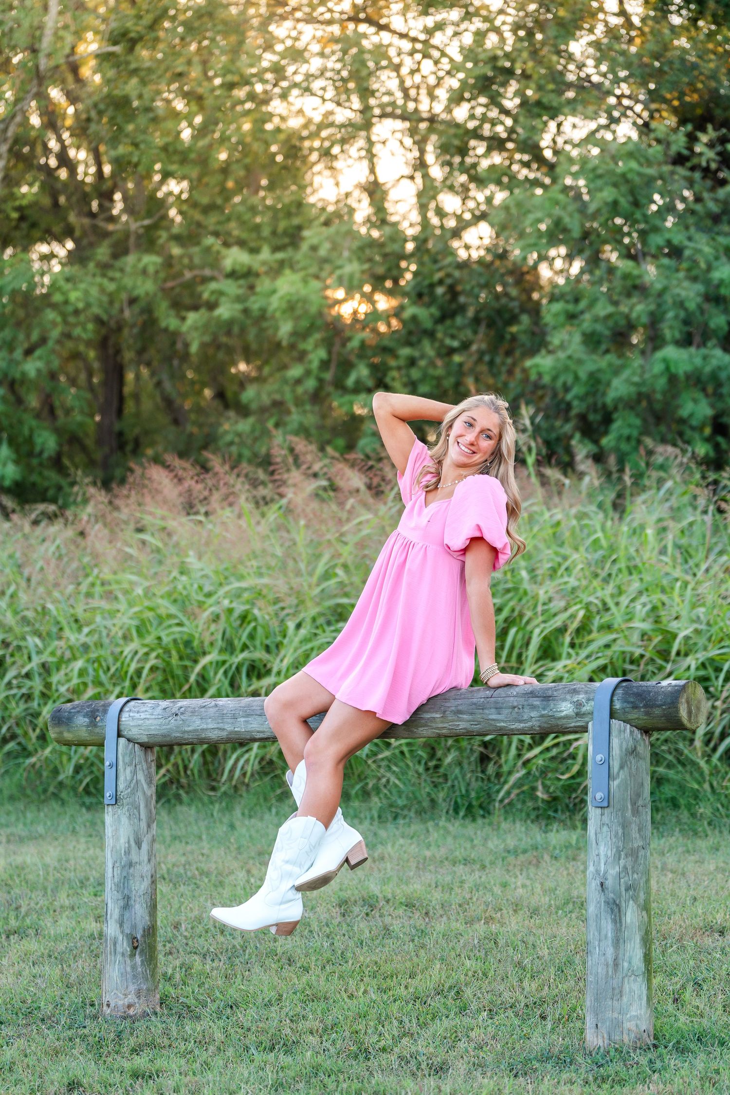 Cheerful blonde woman in pink dress and white cowboy boots sitting on wooden fence rail outdoors.