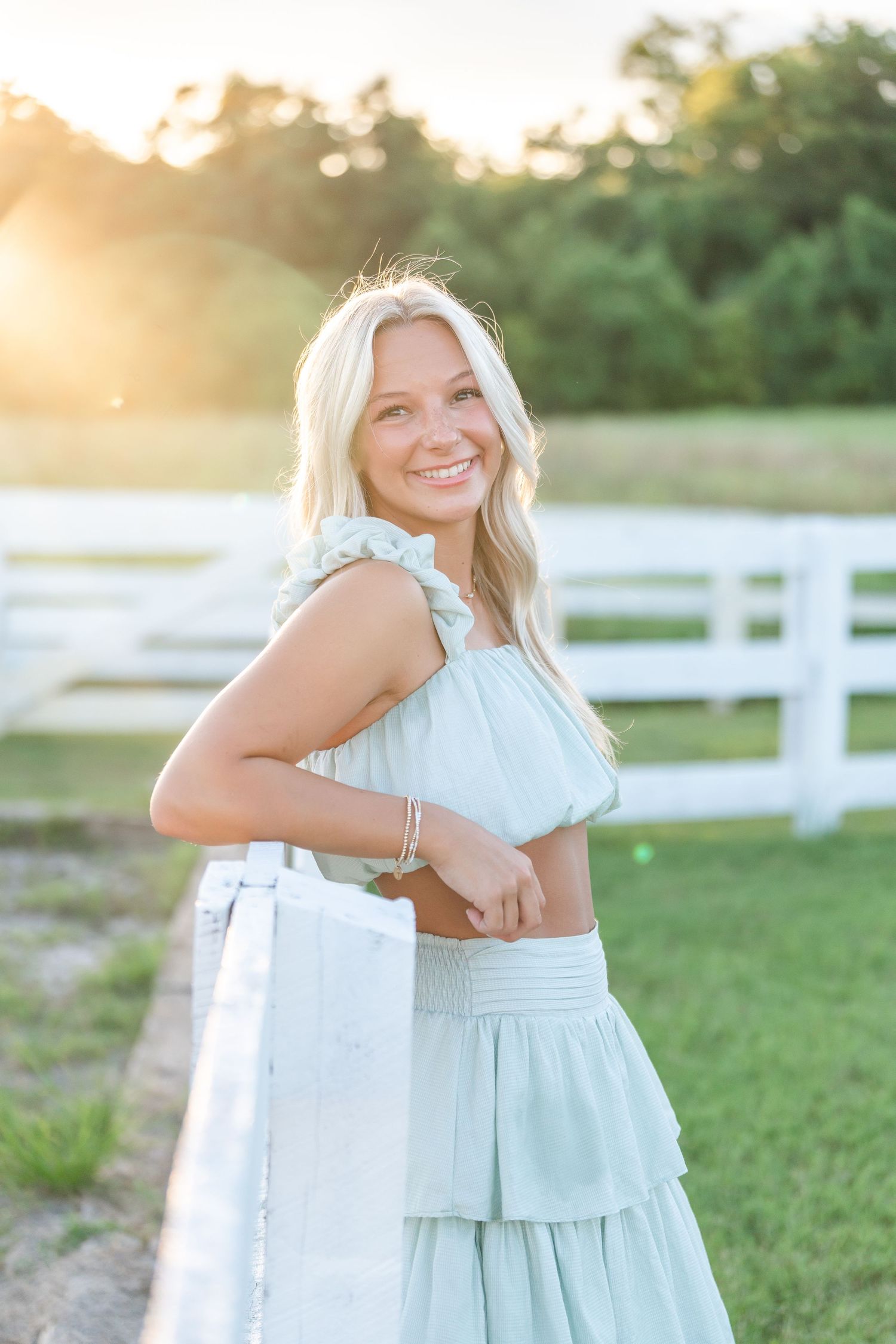 Smiling blonde woman in mint green crop top and tiered skirt posing by white fence at sunset.
