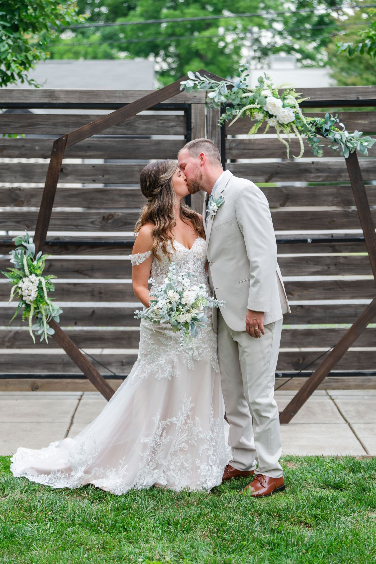 Bride and groom sharing first kiss at altar before rustic wooden hexagon arch with white floral decor.
