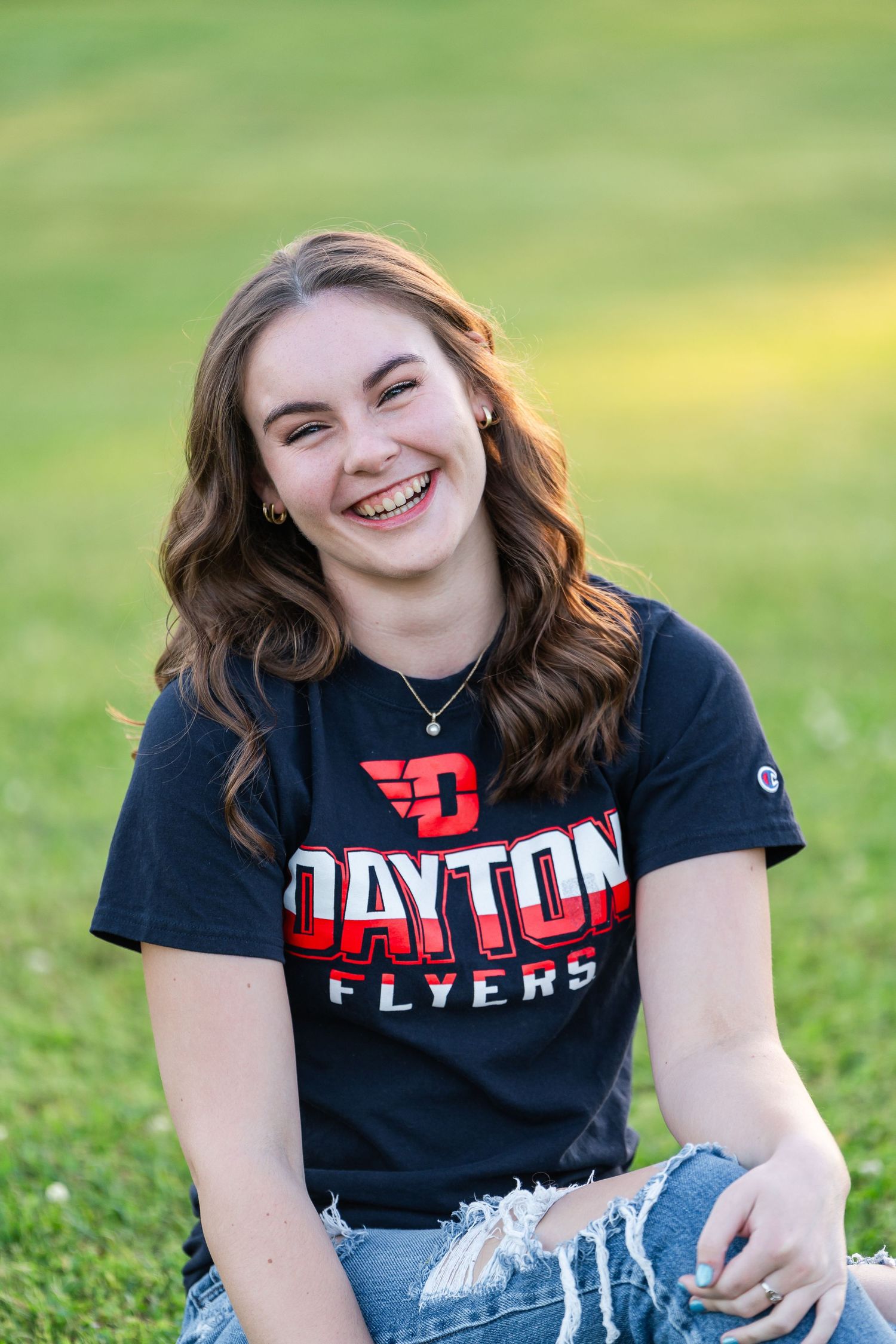 Laughing young woman in Dayton Flyers shirt sitting on green grass in warm natural light.