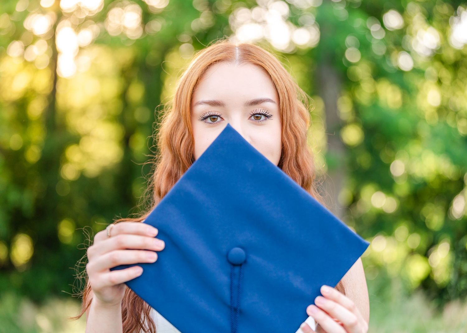 Redheaded graduate playfully hiding her face behind a blue mortarboard cap outdoors with a soft bokeh background.