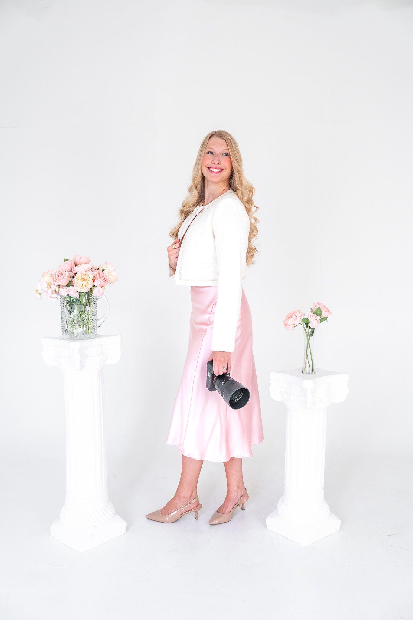 Blonde female photographer in pink satin skirt and white jacket holding camera in studio.