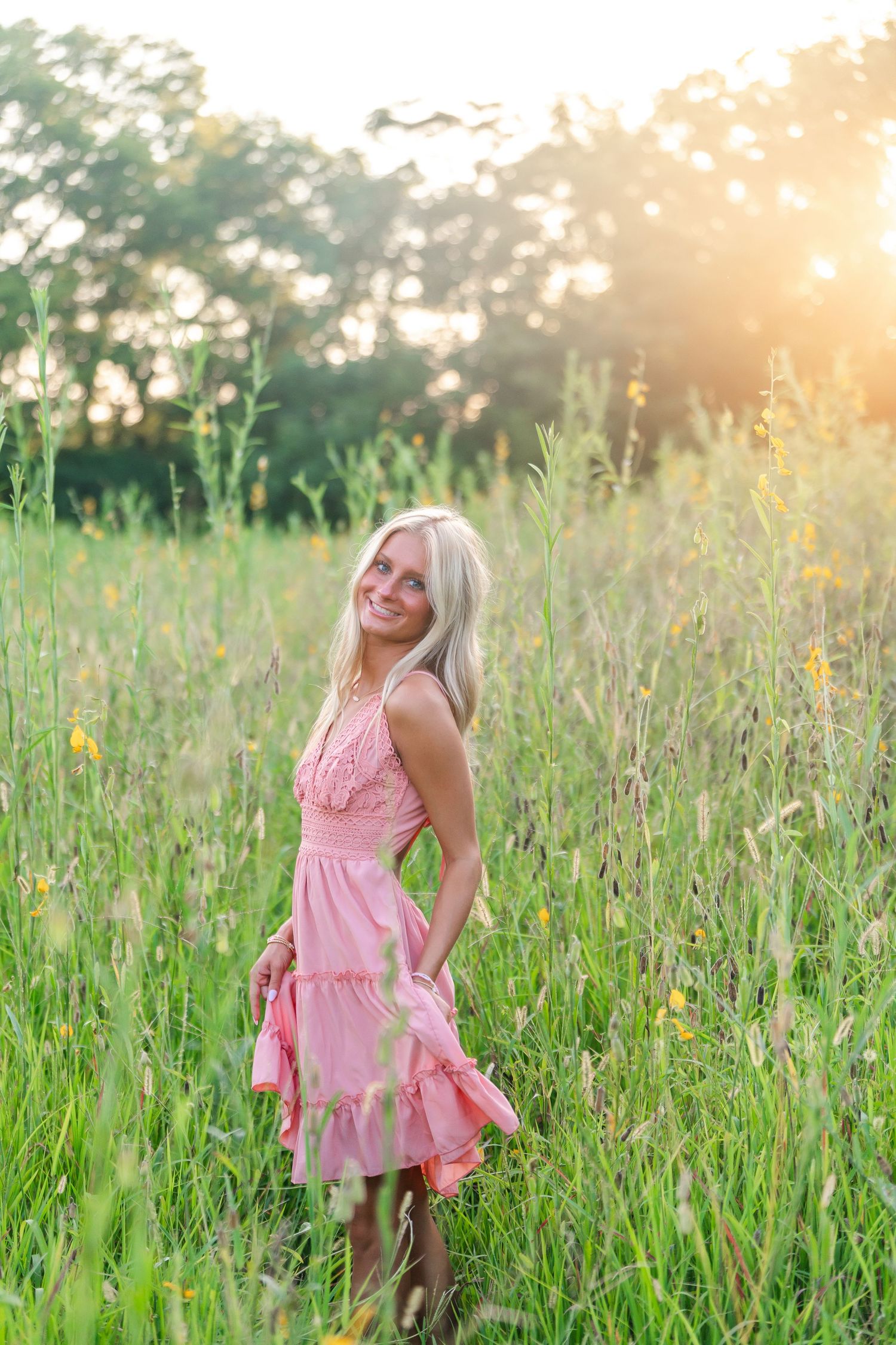 Smiling blonde woman in pink dress standing in sunny green meadow with wildflowers and golden backlight.