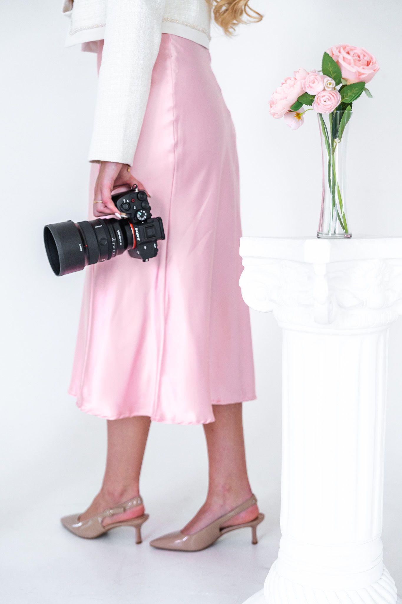 Close-up of woman in pink satin skirt holding professional camera beside pink roses in vase.