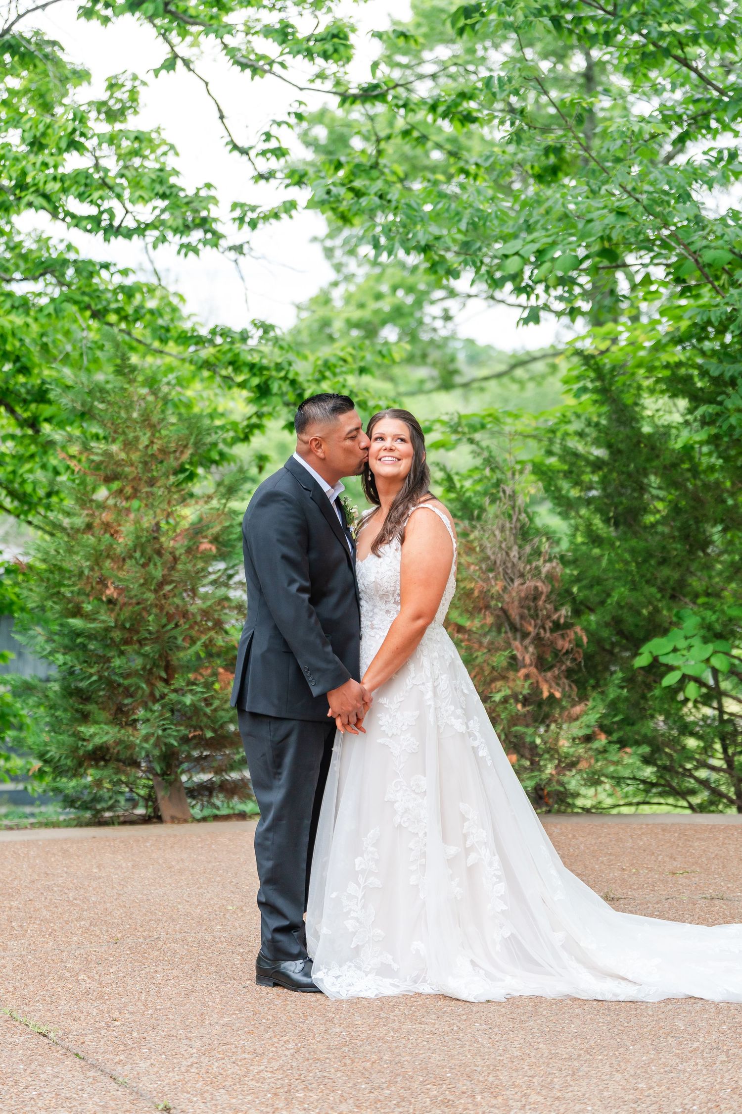 Bride and groom holding hands and sharing a kiss outdoors surrounded by green trees.