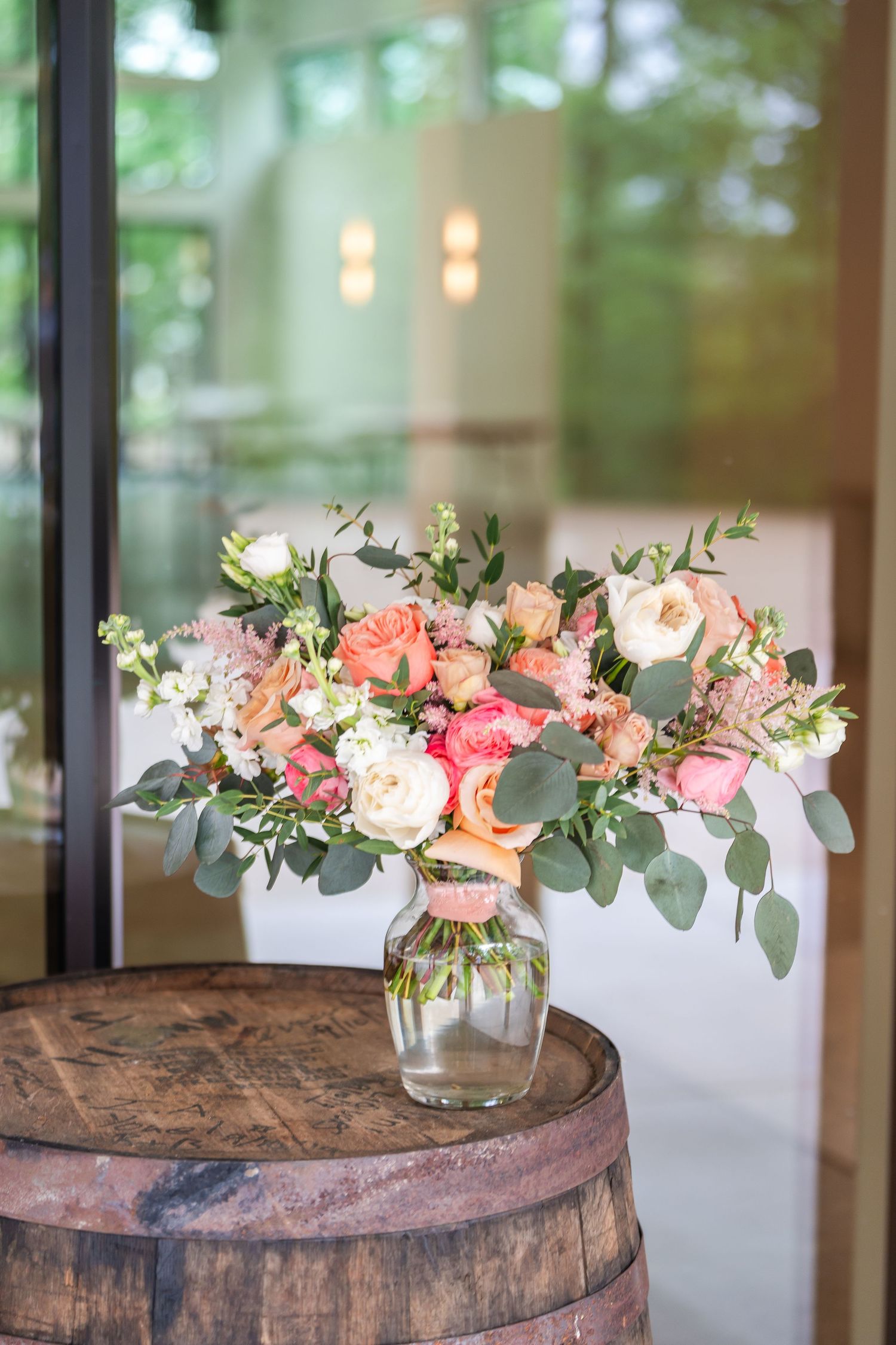 Lush bridal bouquet of coral, peach, and white roses with eucalyptus in a glass vase on a rustic wooden barrel.
